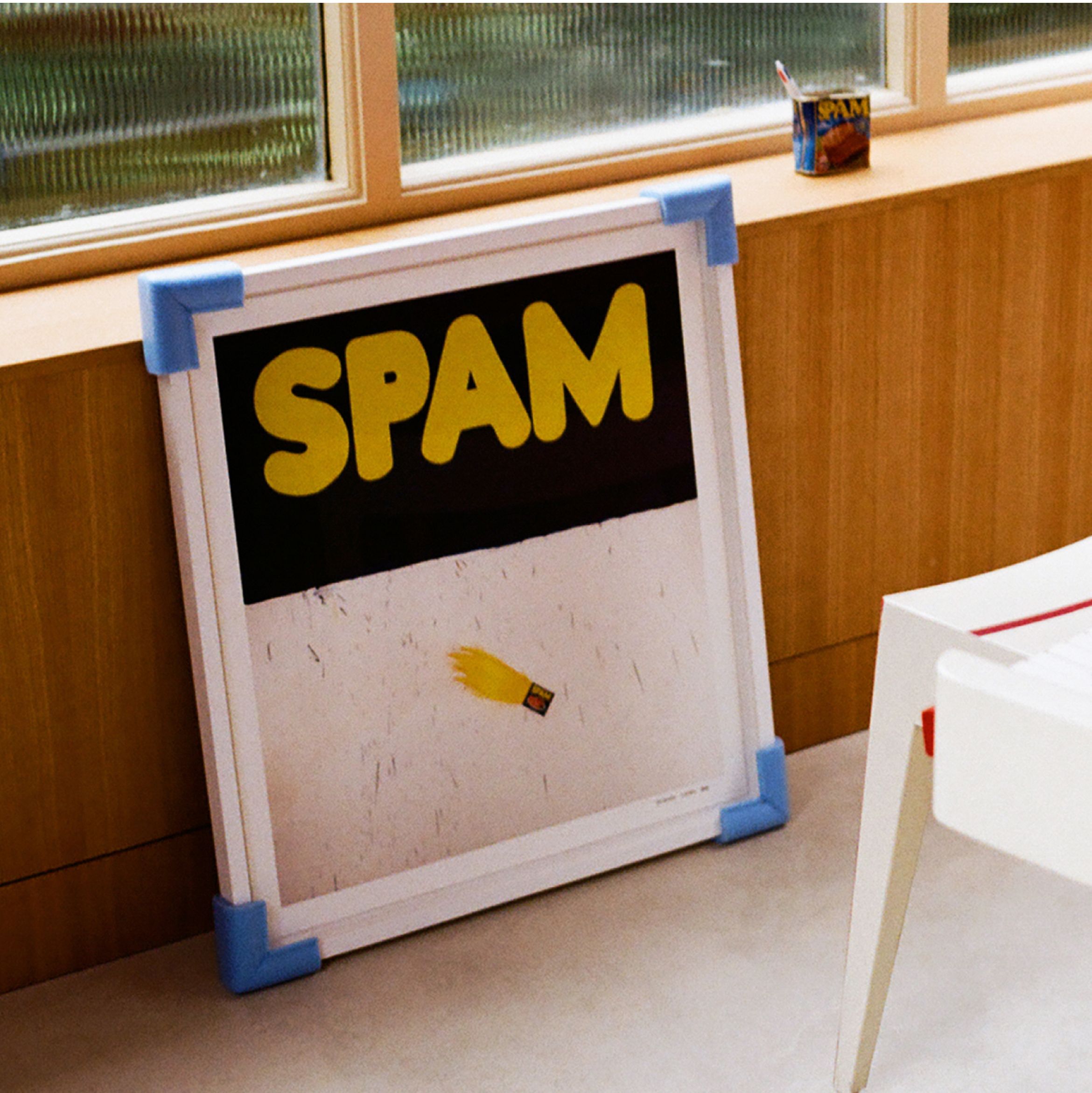 Framed SPAM poster with blue corner protectors, leaning against a wood-paneled wall near a window, with a SPAM can on the ledge.