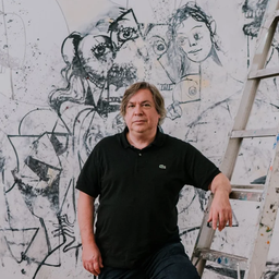 George Condo stands infront of work in progress painting, arm placed on stepladder wearing a black shirt