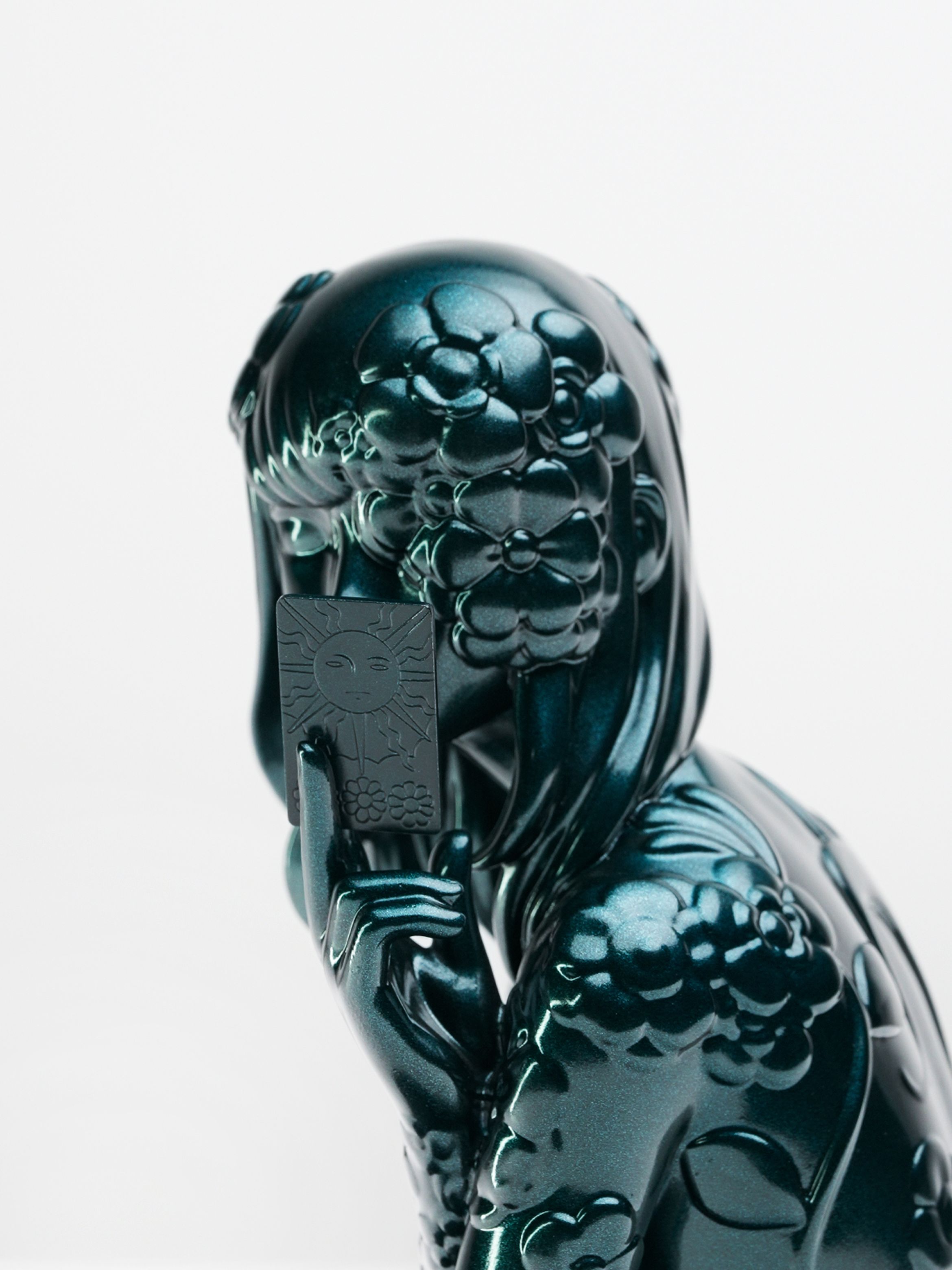 Close-up of a sculpture of a person with stylised hair and a flower detail, featuring a reflective, dark teal surface.
