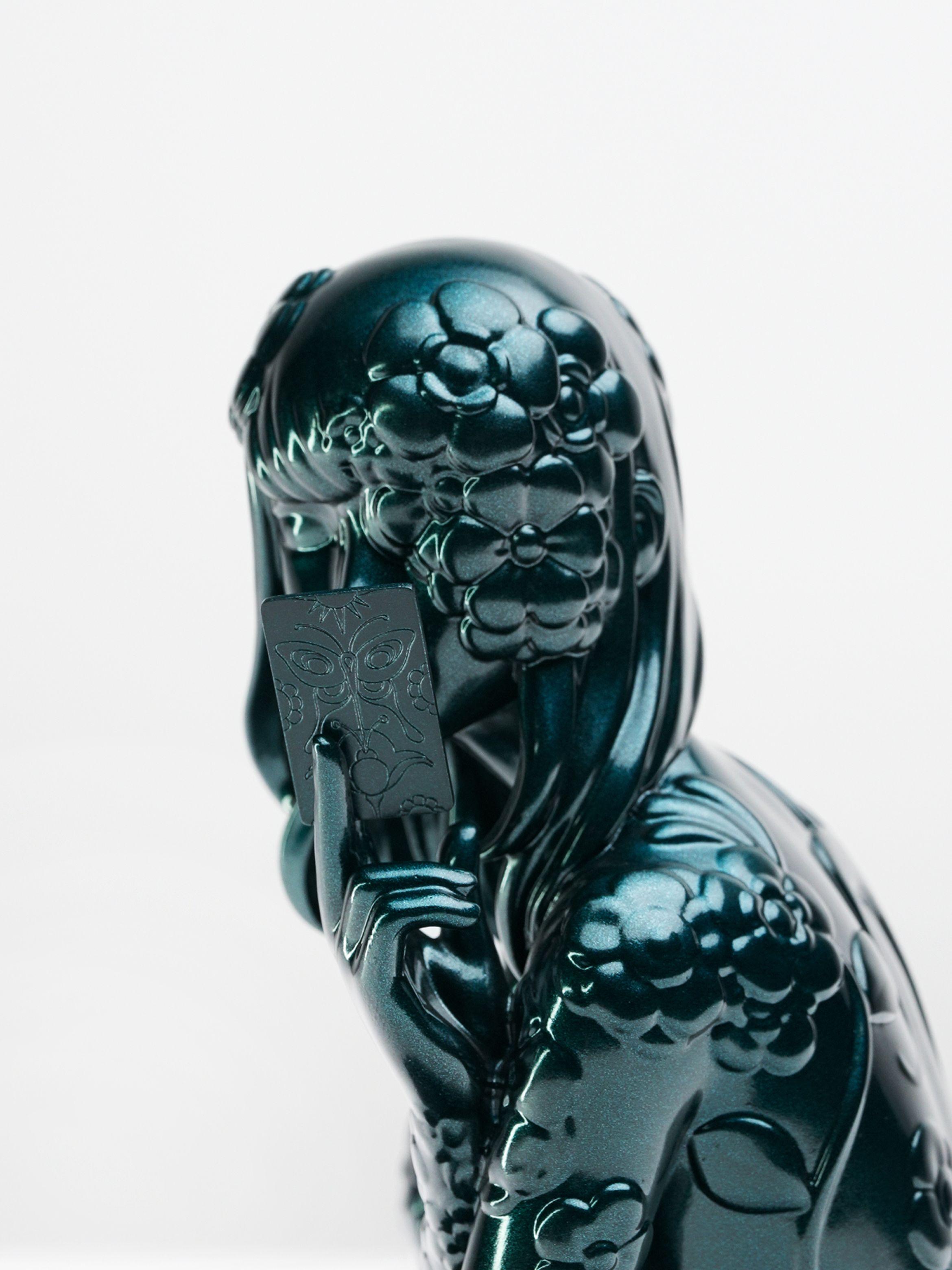 Close-up of a sculpture of a person with stylised hair and a flower detail, featuring a reflective, dark teal surface.
