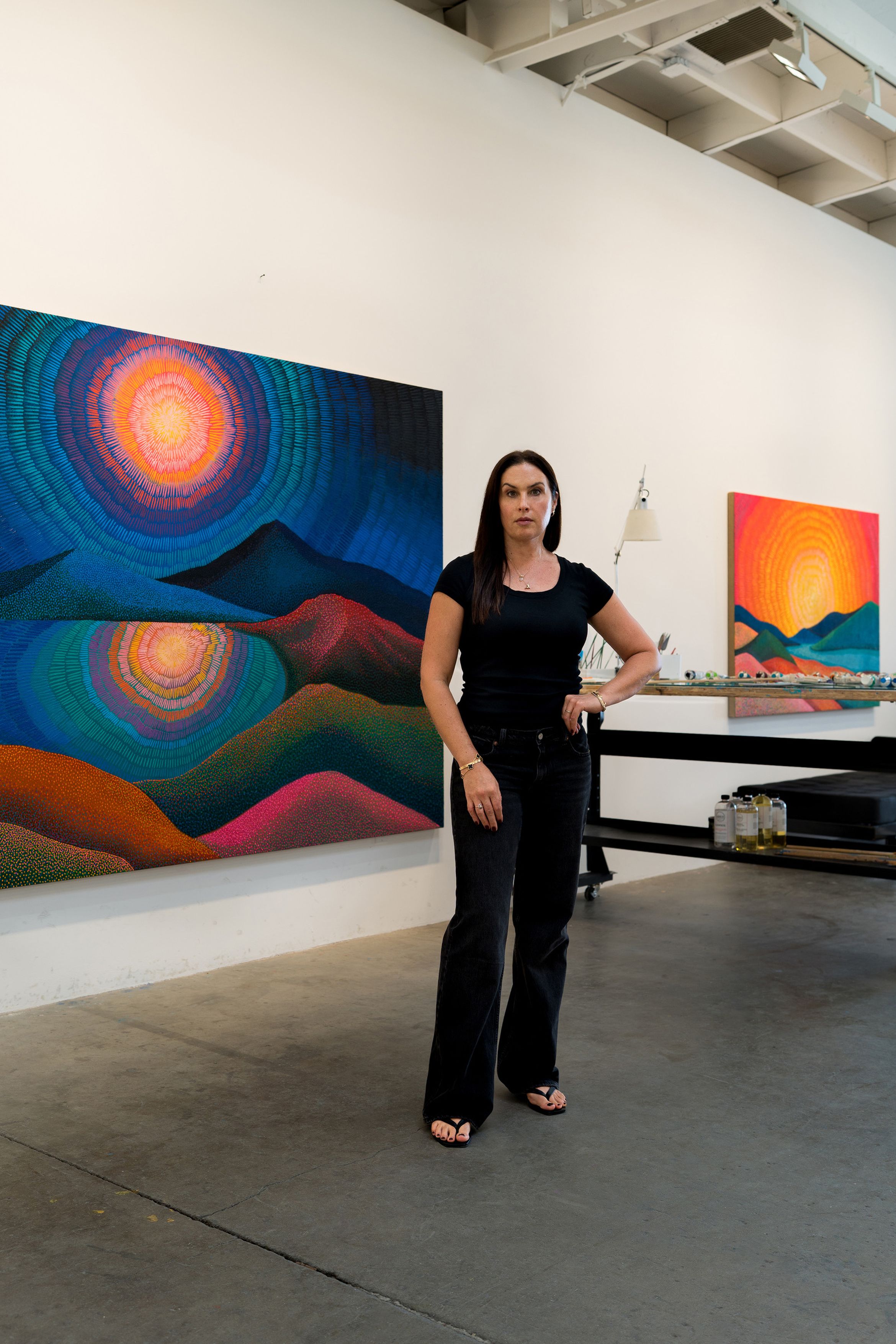 A woman in a black outfit stands in an art studio, surrounded by vibrant, colorful landscape paintings on the walls.