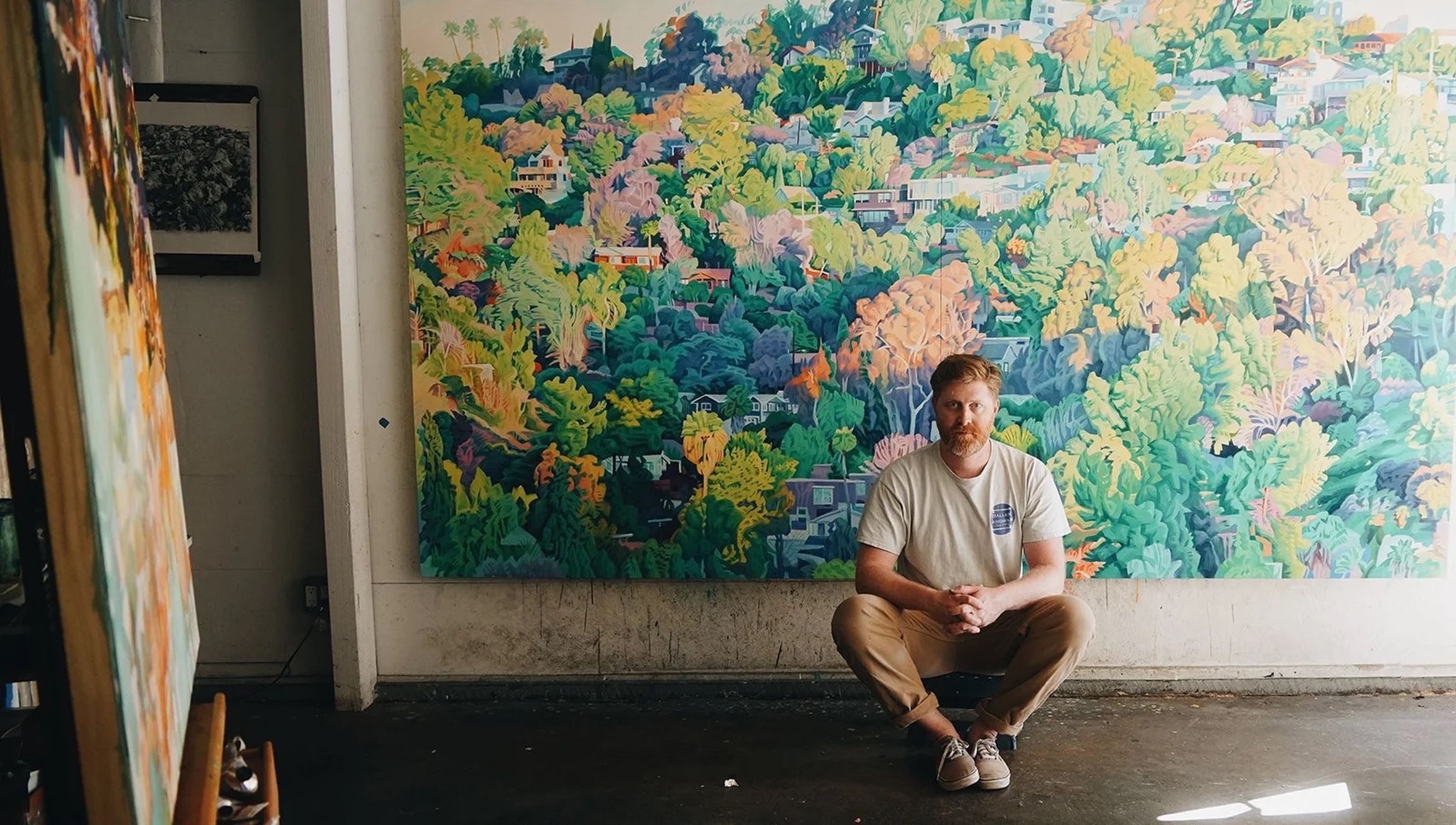 A person with red hair sits in front of a large colorful painting depicting a vibrant, detailed landscape with foliage and houses.