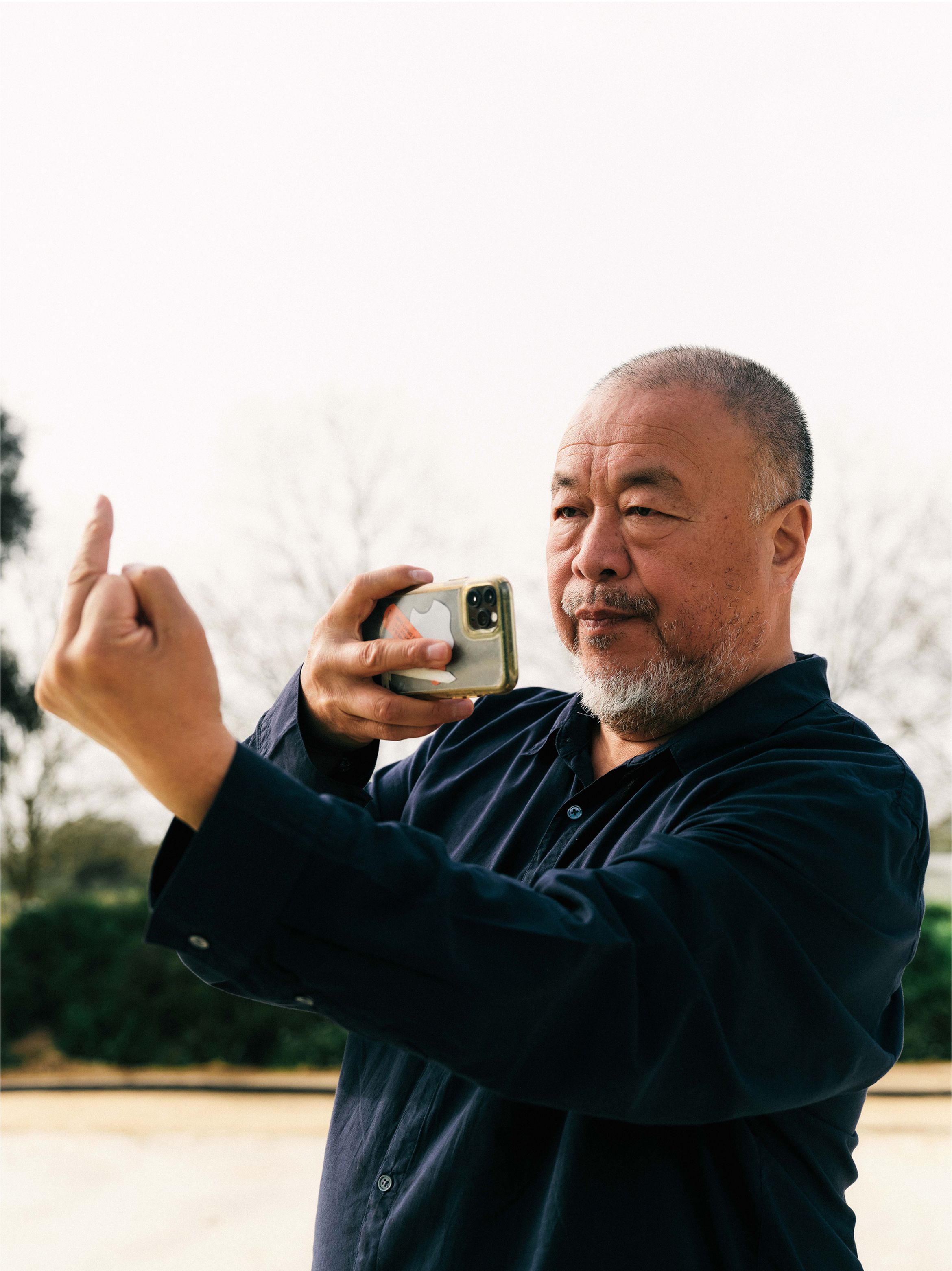A man in a dark shirt takes a selfie with a smartphone while raising his middle finger, standing outdoors.