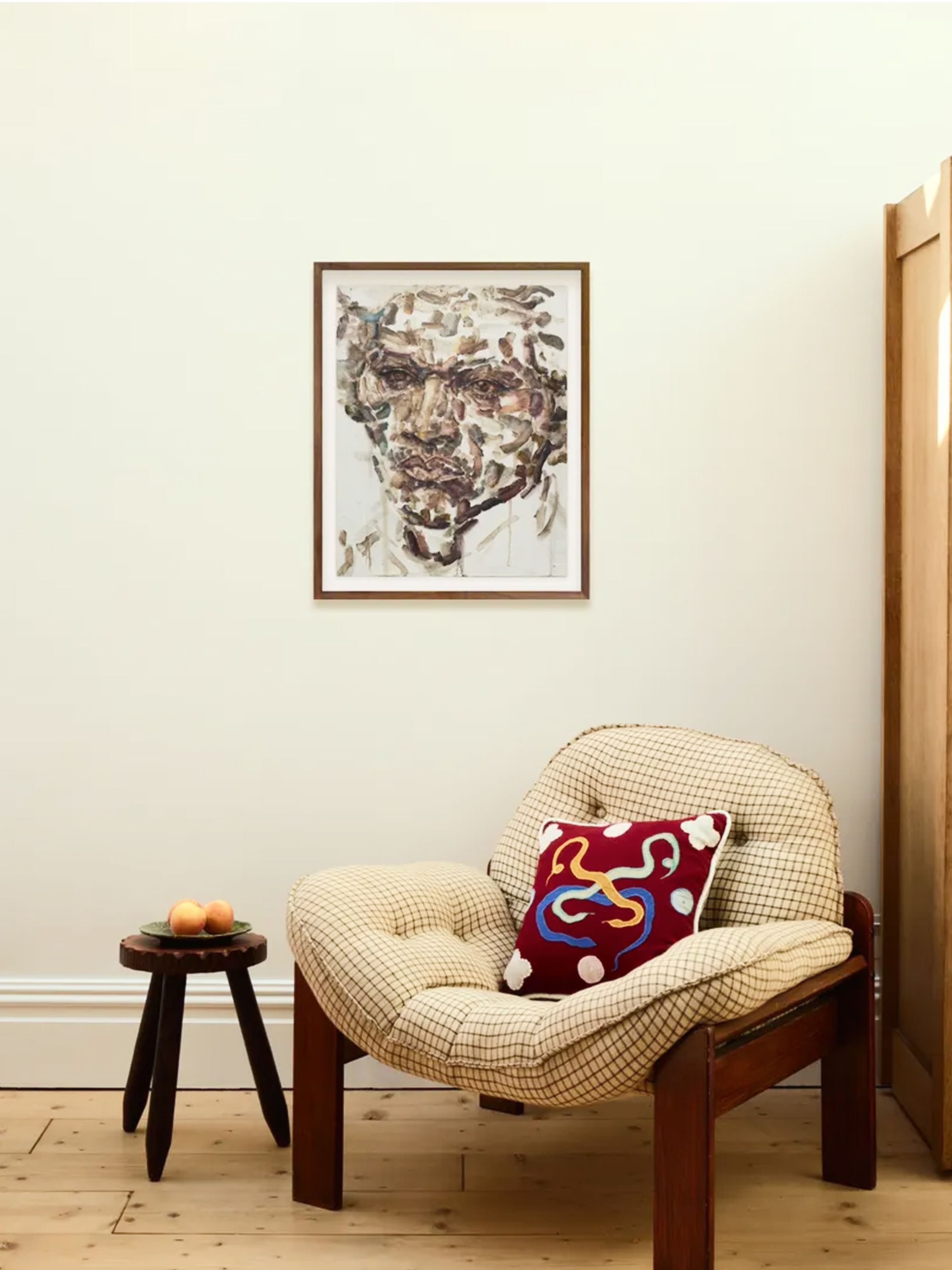 Cozy room with a cushioned chair, colorful pillow, small wooden table with fruit, and abstract face artwork on a cream wall.