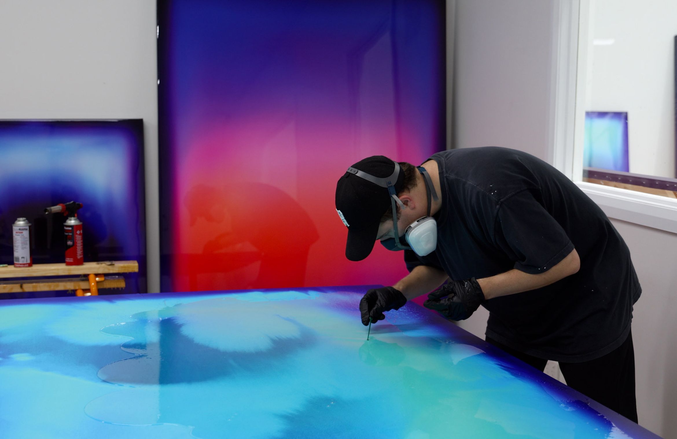 Artist using a tool to create abstract art on a colorful, gradient canvas, wearing a cap, mask, and gloves in a studio setting.