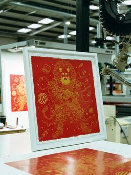 A framed red and gold artwork depicting an intricate, mythical figure is displayed on a table in an art studio.