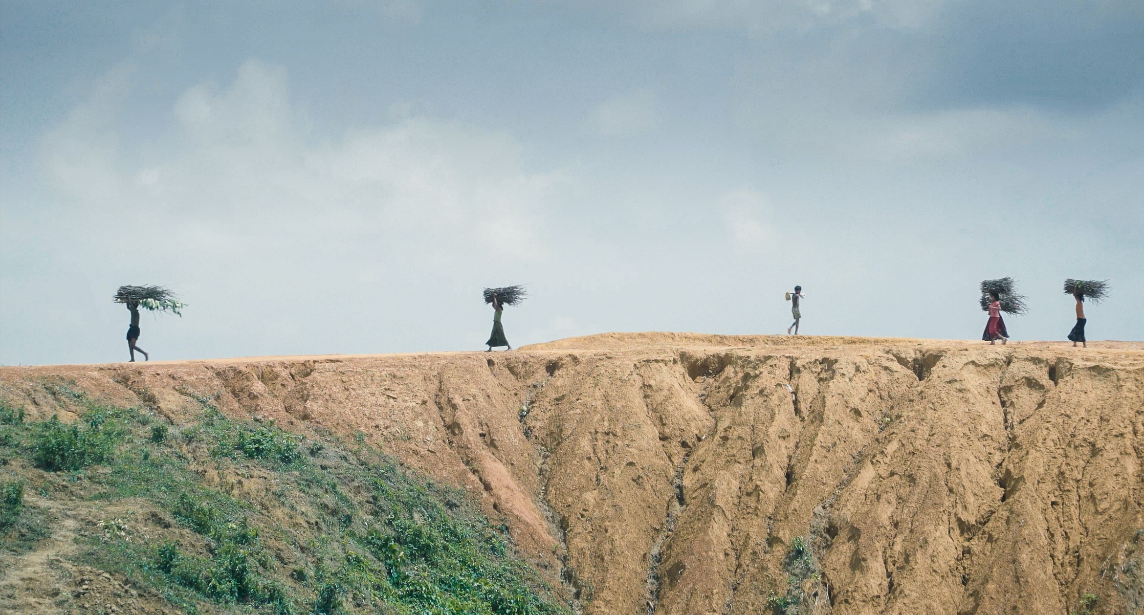 Three people walk along a ridge carrying bundles of sticks, set against a cloudy sky and rugged landscape. Three people walk along a ridge carrying bundles of sticks, set against a cloudy sky and rugged landscape.