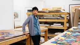 artist in paint-splattered denim shirt, leaning on a table on which a row of colourful prints are laid artist in paint-splattered denim shirt, leaning on a table on which a row of colourful prints are laid