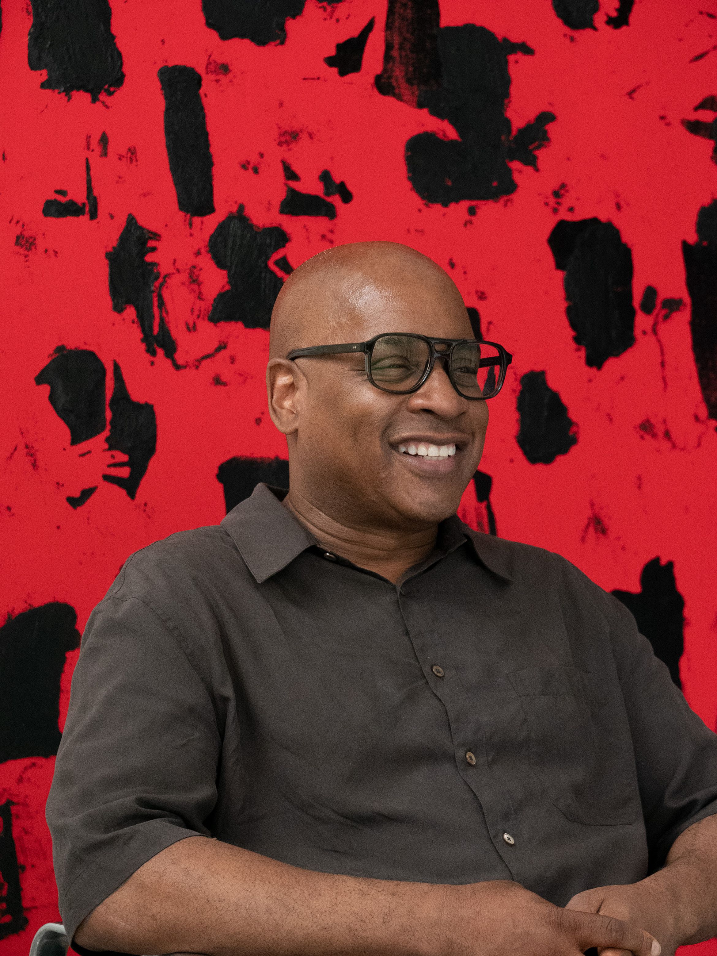 A smiling person (Artist Glenn Ligon) wearing glasses and a dark shirt sits in front of a bold red and black abstract background.