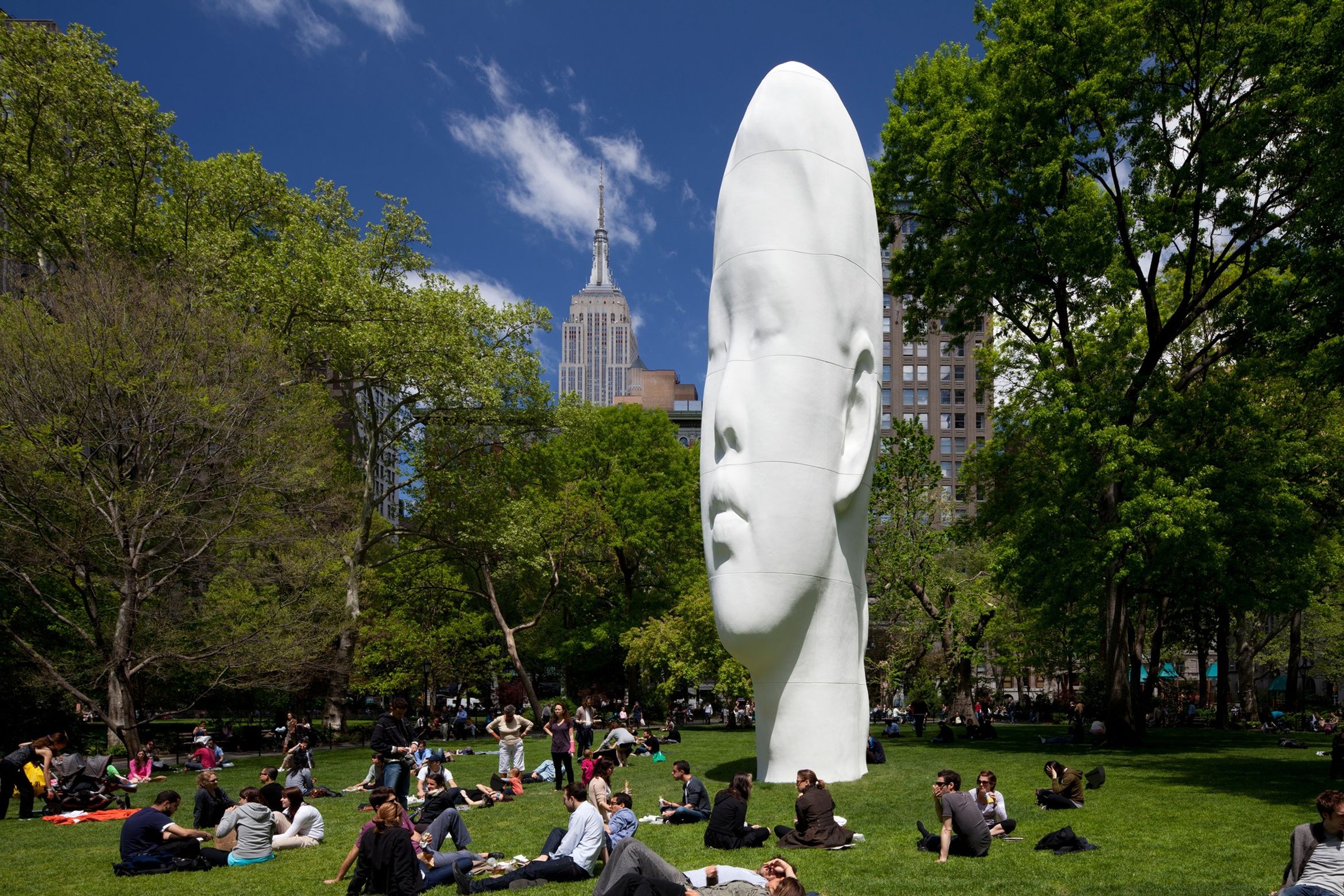 Large white head sculpture in a park with people relaxing on the grass; tall buildings in the background, including the Empire State Building.