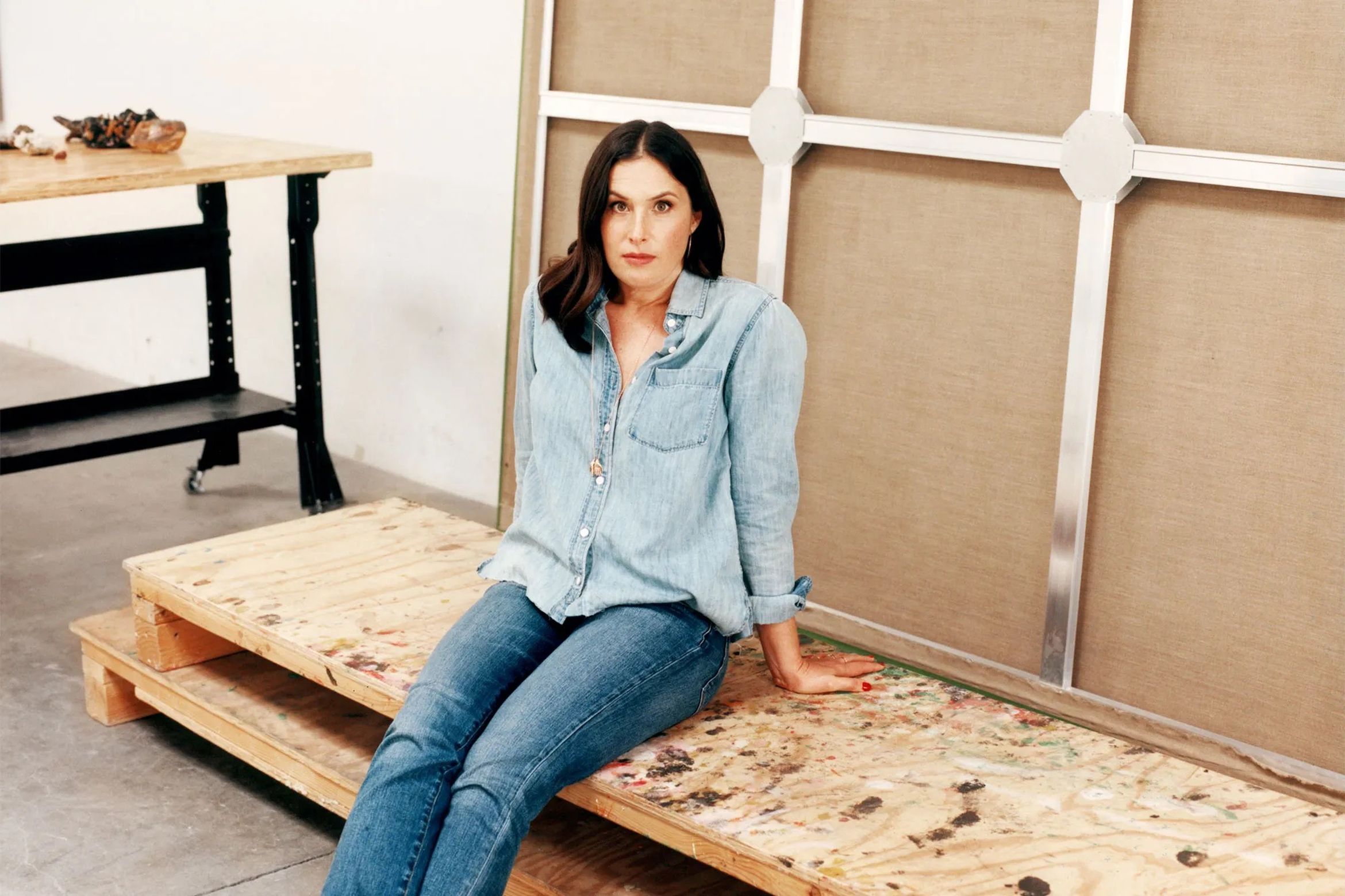 A woman in a denim shirt and jeans sits on a wooden platform next to a large canvas frame in an art studio.