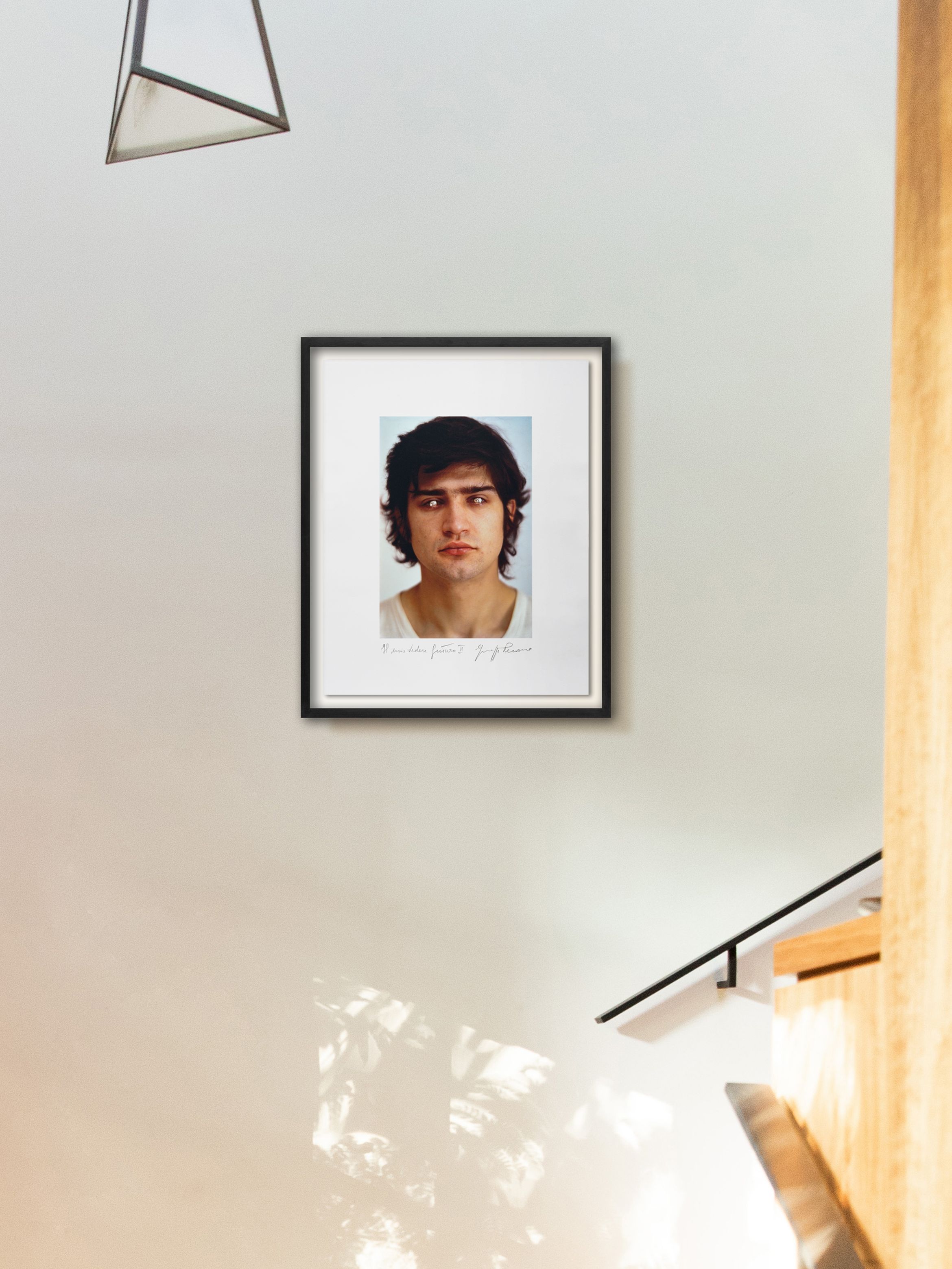 Framed portrait on a light wall beside a staircase, with geometric pendant above and soft light reflections below.
