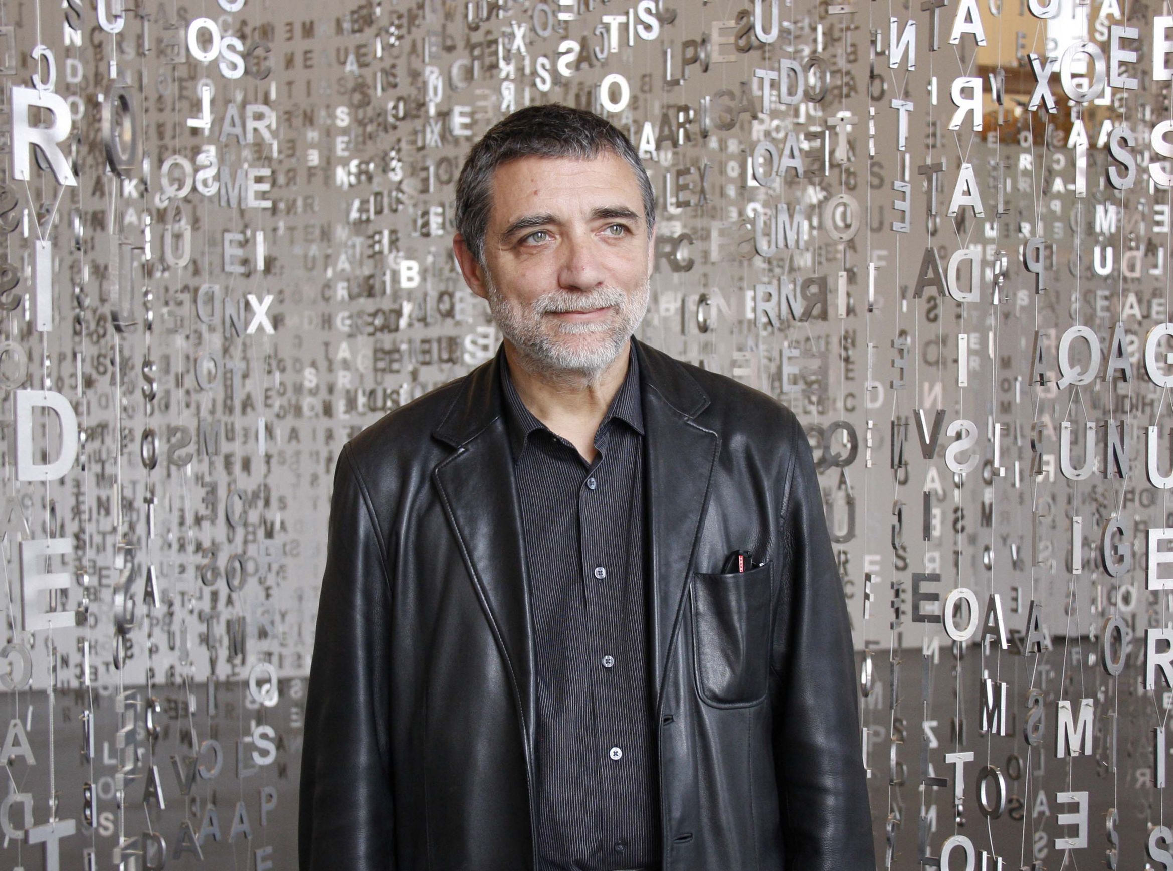 Man in a black leather jacket stands in front of a backdrop of suspended letters, creating a textured and artistic background.