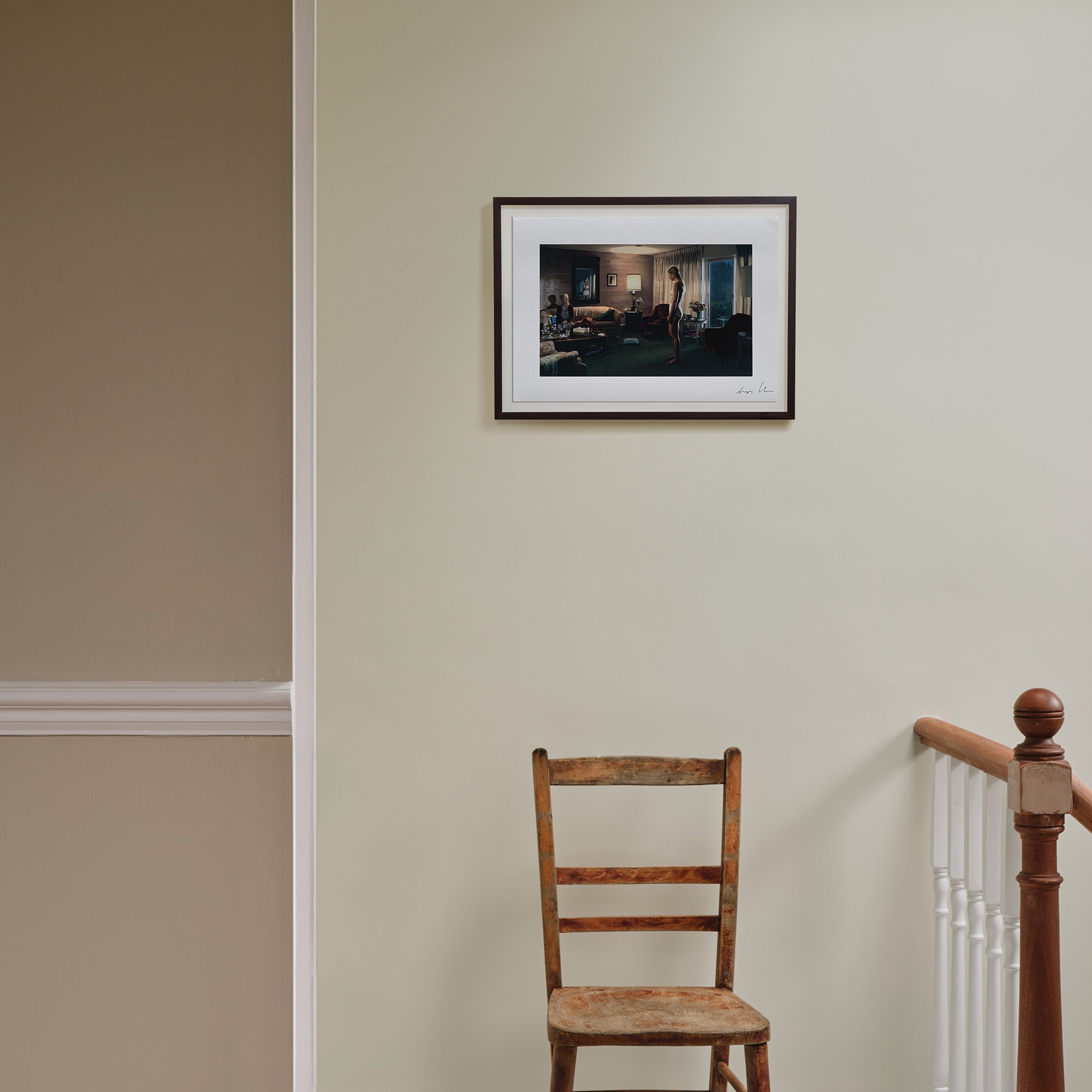 A wooden chair sits beneath a framed photo on a pale green wall, next to a staircase with a wooden railing.