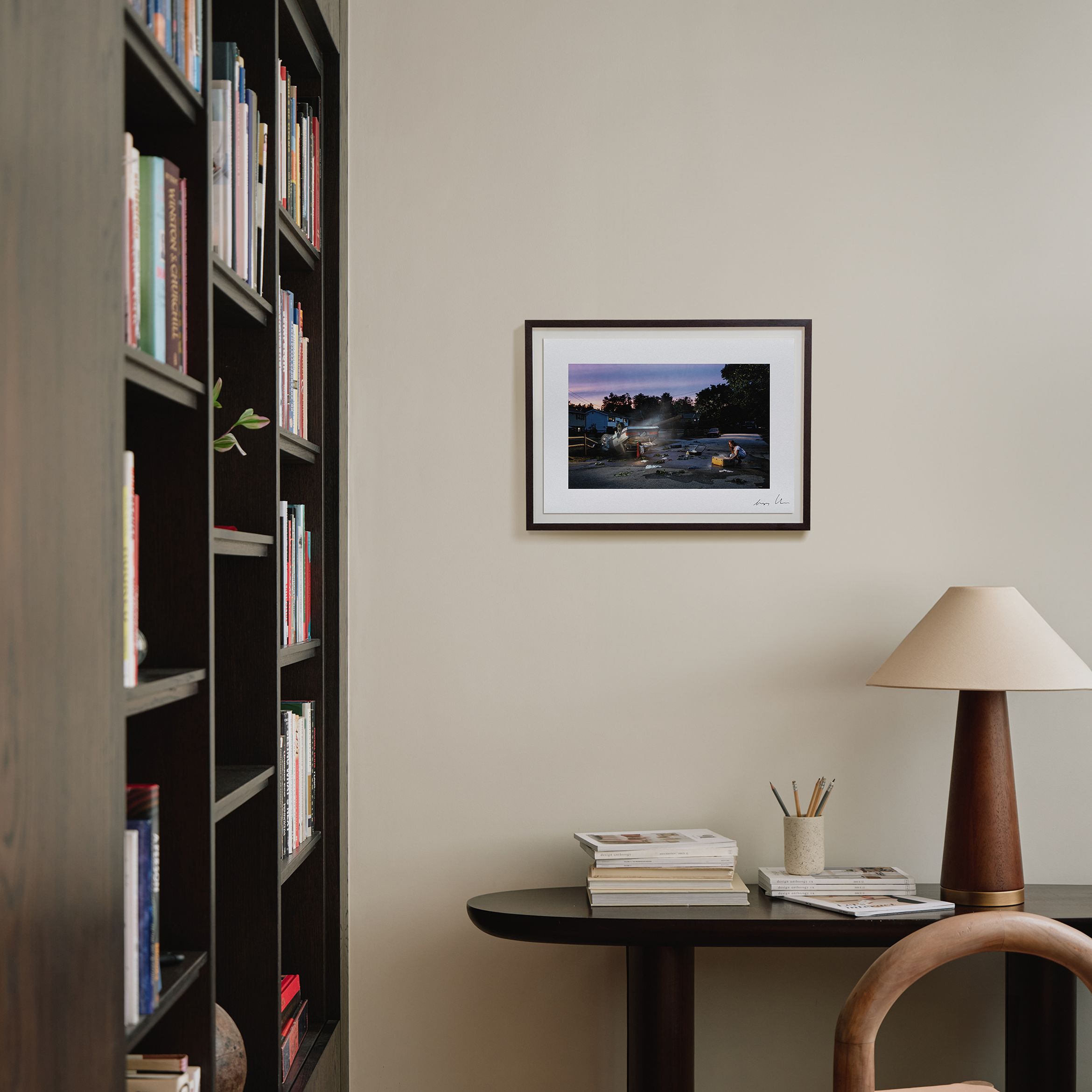 Bookshelf and desk with a lamp, books, and pencils. A framed photo of a sunset scene hangs on the beige wall above the desk.
