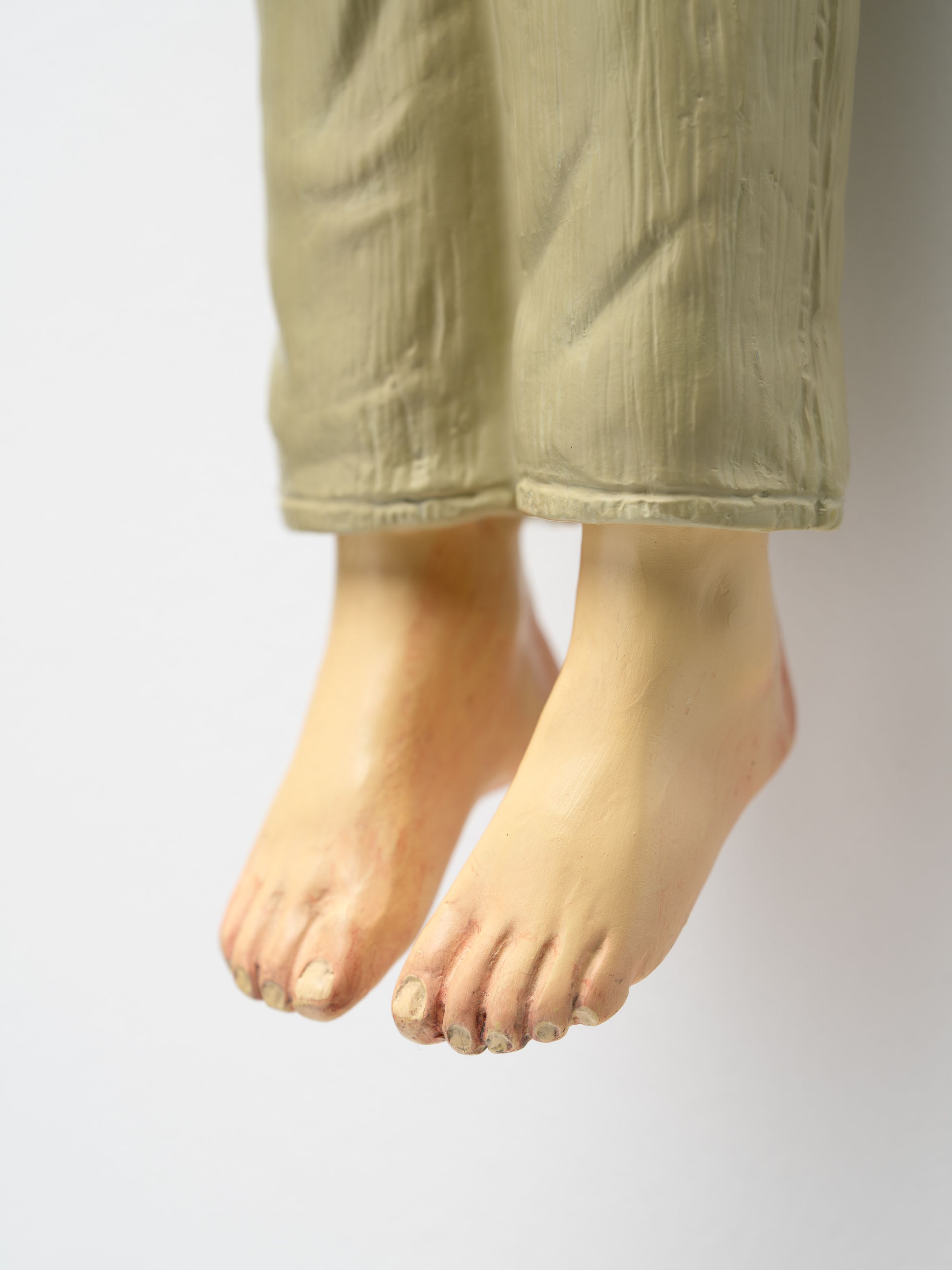 Close-up of a sculpture depicting human feet and lower legs wearing beige pants, against a plain white background.