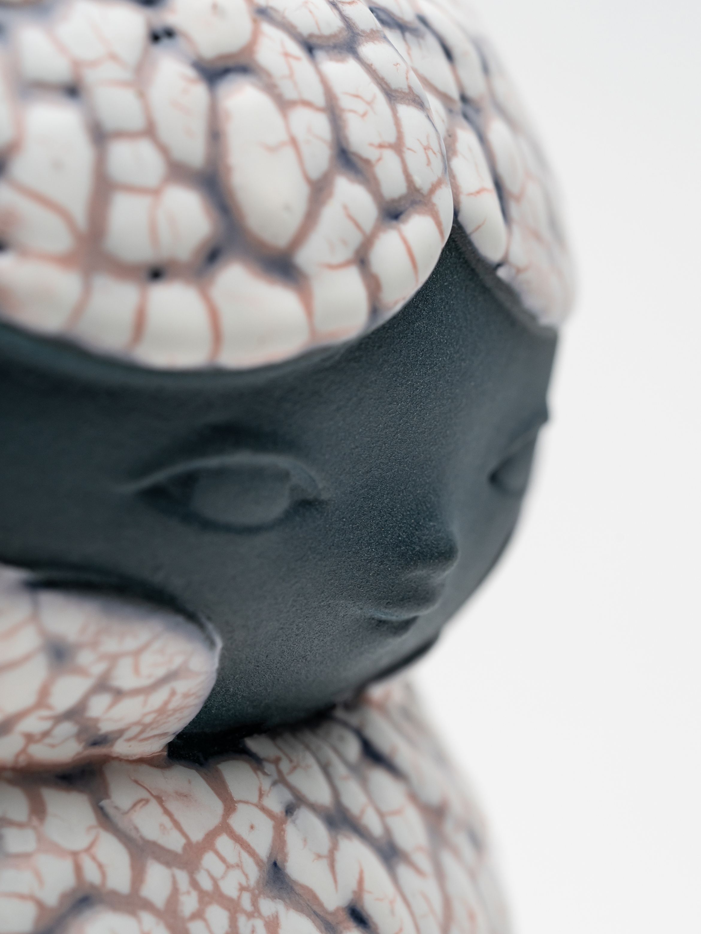 Close-up of a ceramic sculpture with a dark face and textured, cracked white and pink surface resembling hair or scales.