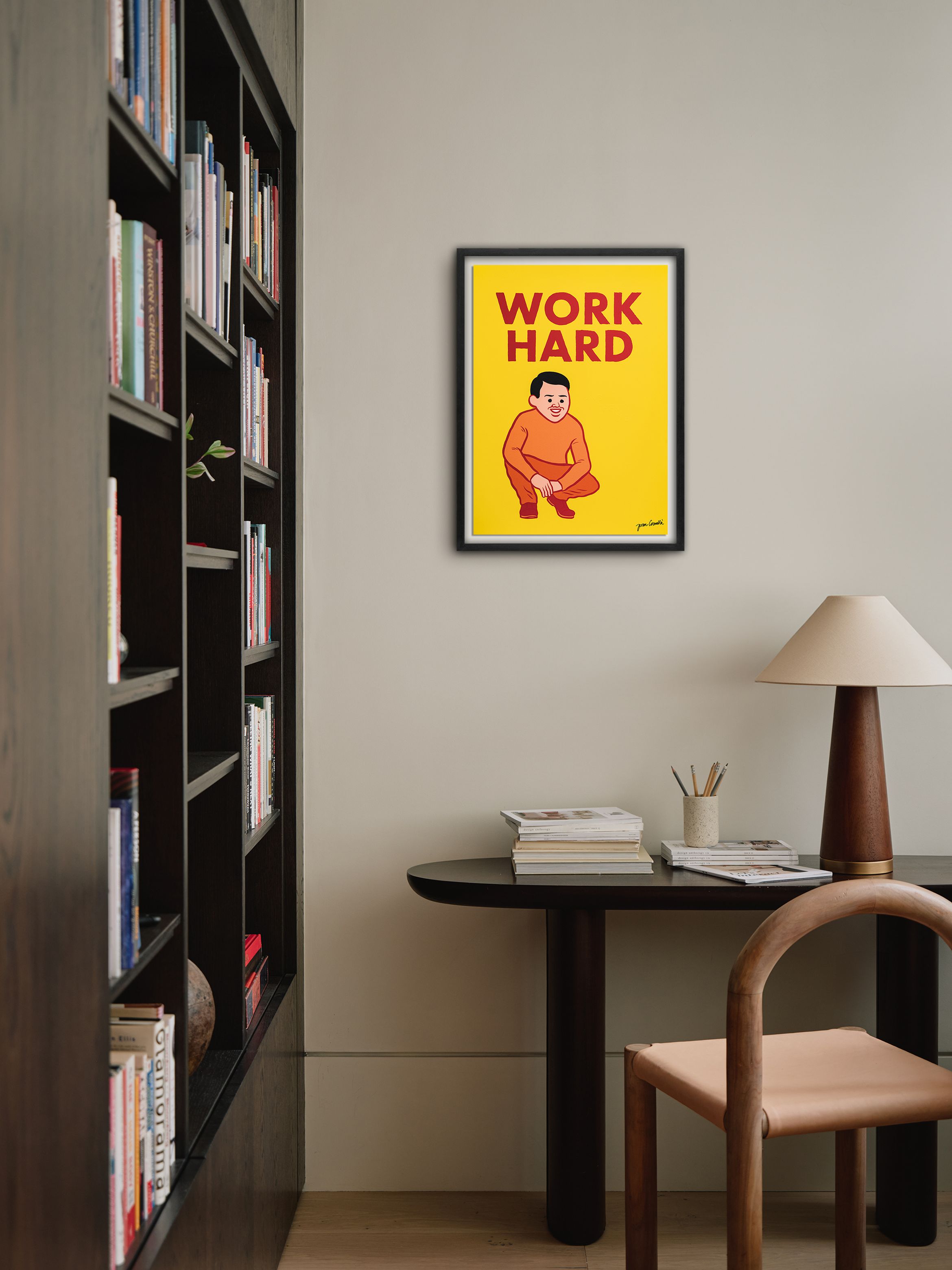 Home office with a wooden desk, chair, and lamp. Bookshelf on the left. A framed "Work Hard" poster with an illustration hangs on the wall.
