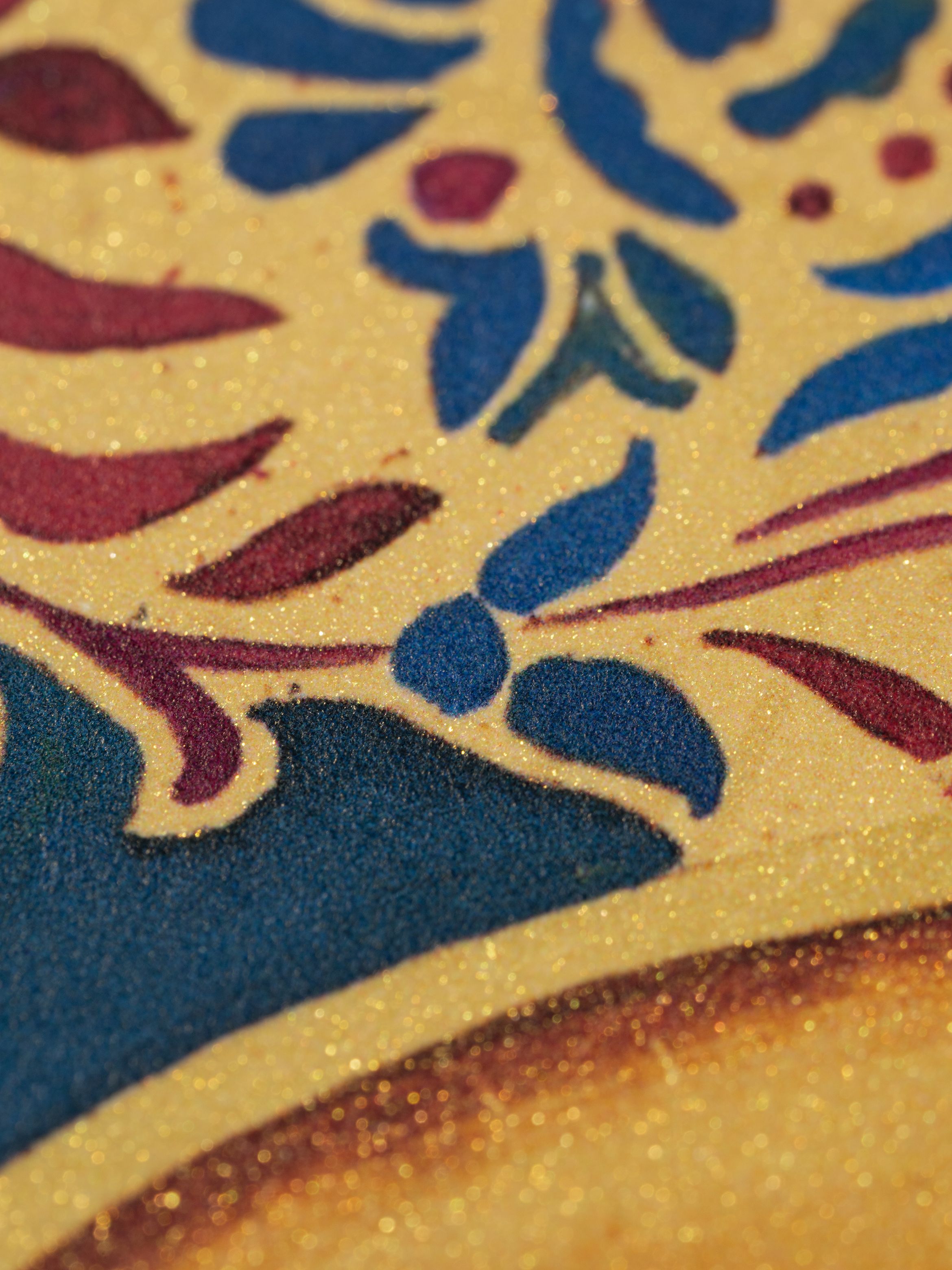 Close-up of a colorful, textured pattern featuring red, blue, and gold floral and abstract designs on a glittery surface.