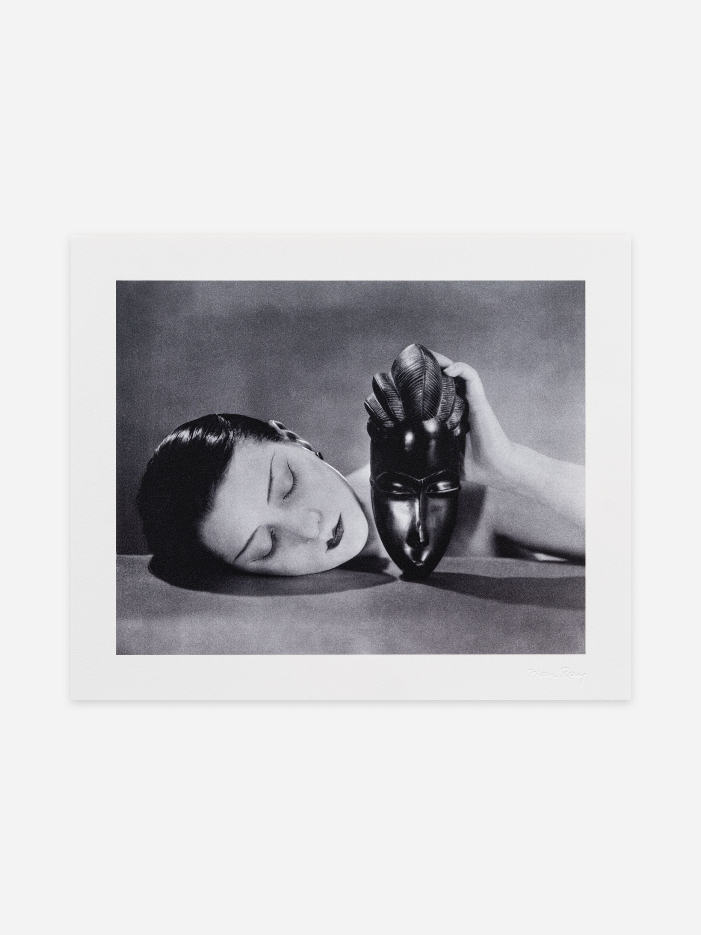 Black-and-white photo of a woman's head resting on a surface, eyes closed, hand holding a dark carved mask beside her face.