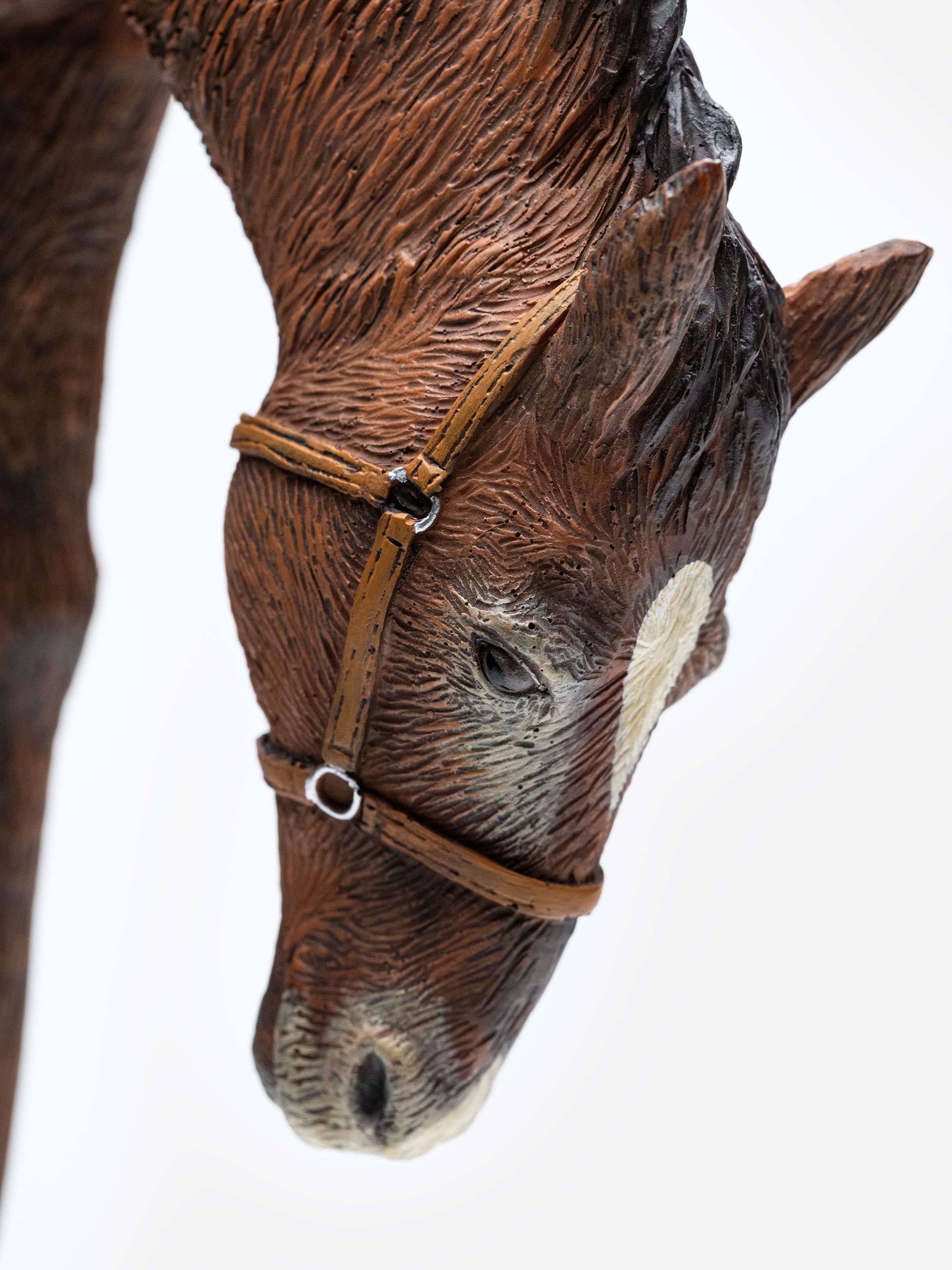 Close-up of a detailed brown horse sculpture with a white blaze and a bridle, facing downward against a plain background.