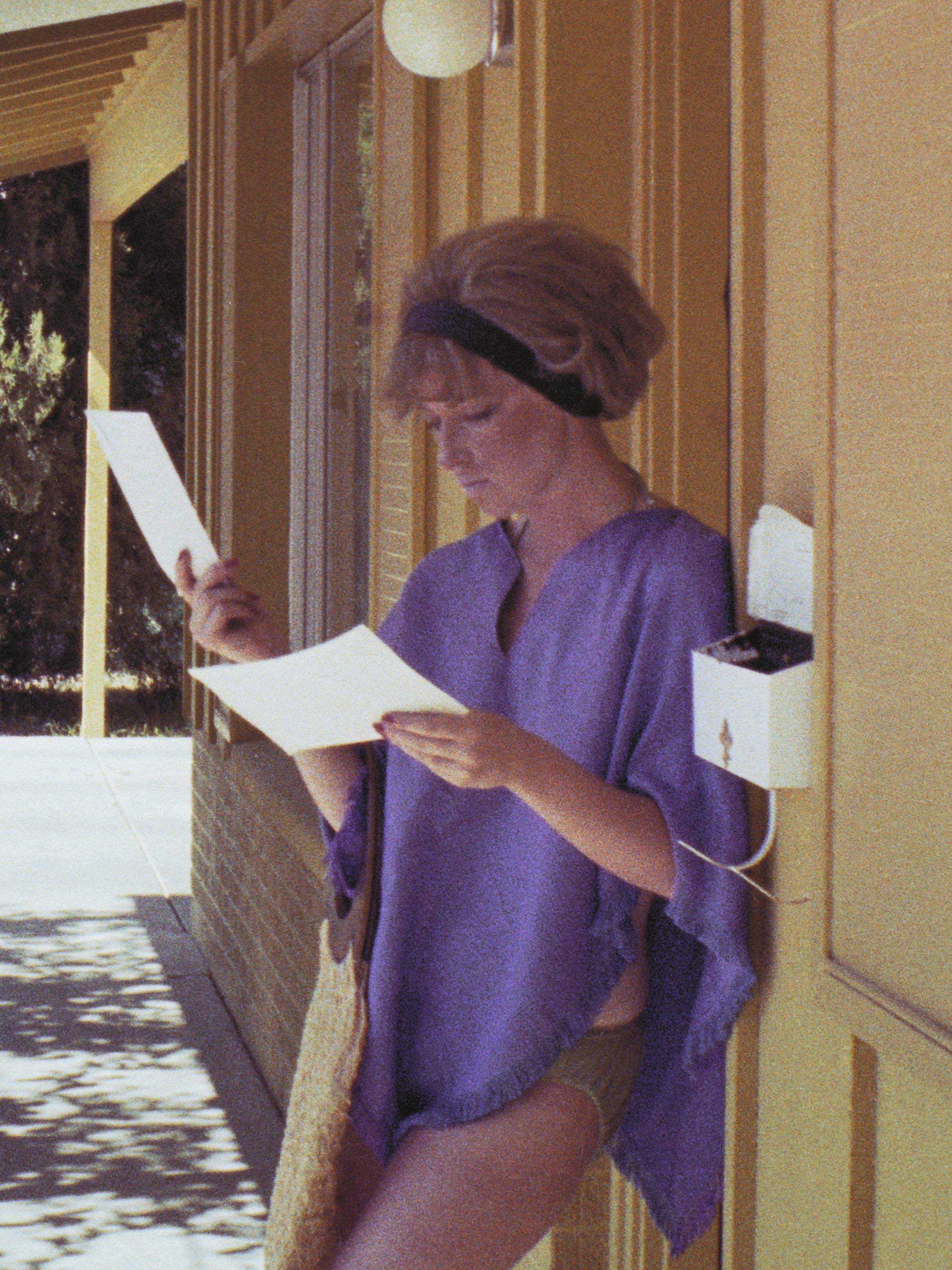 A person in a purple top, holding and reading papers, stands next to a yellow building with a bag hanging from their arm.