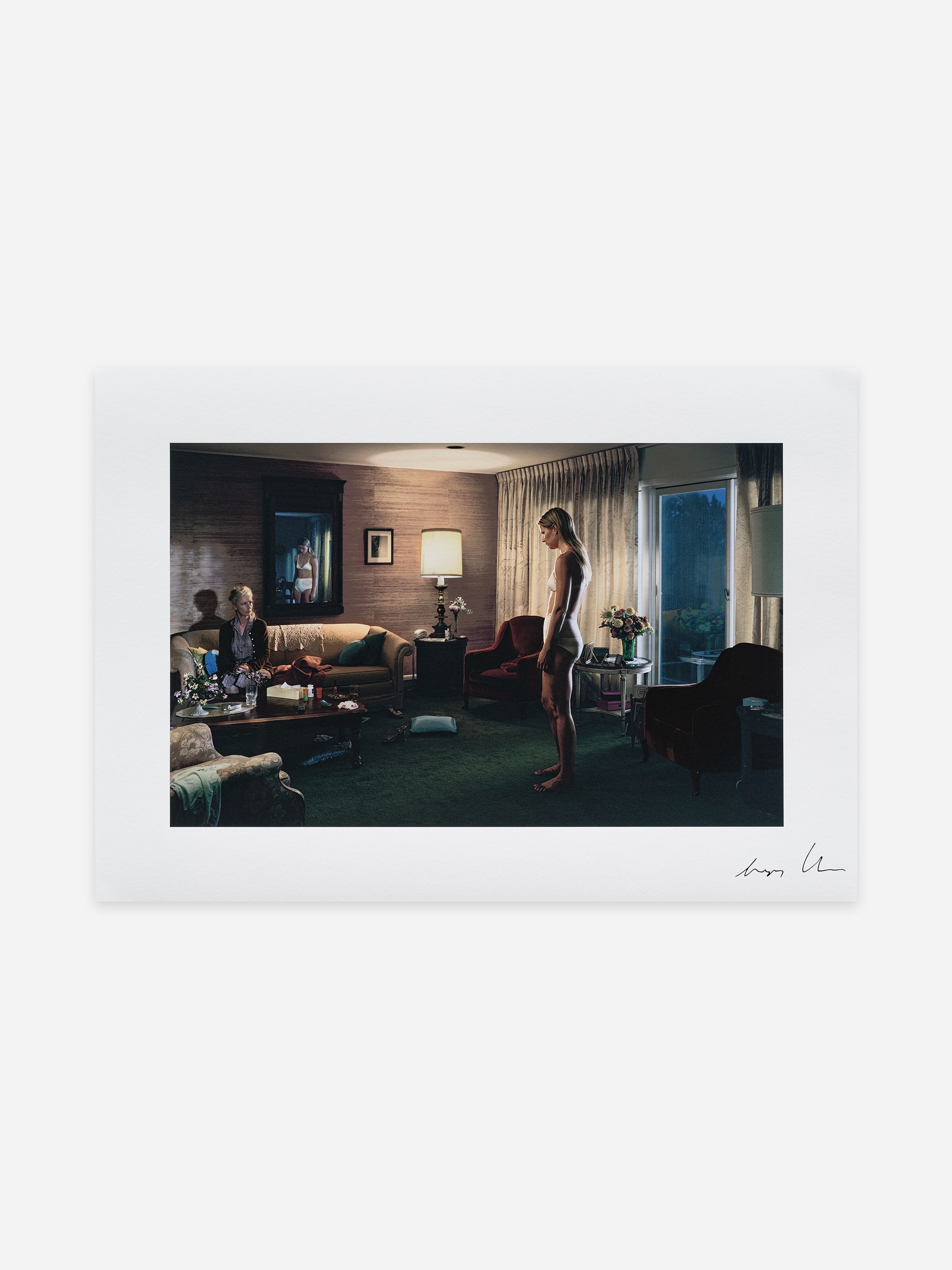 A person stands in a dimly lit living room, wearing a light outfit, while another sits on a sofa. The room has a warm ambiance with soft lighting.