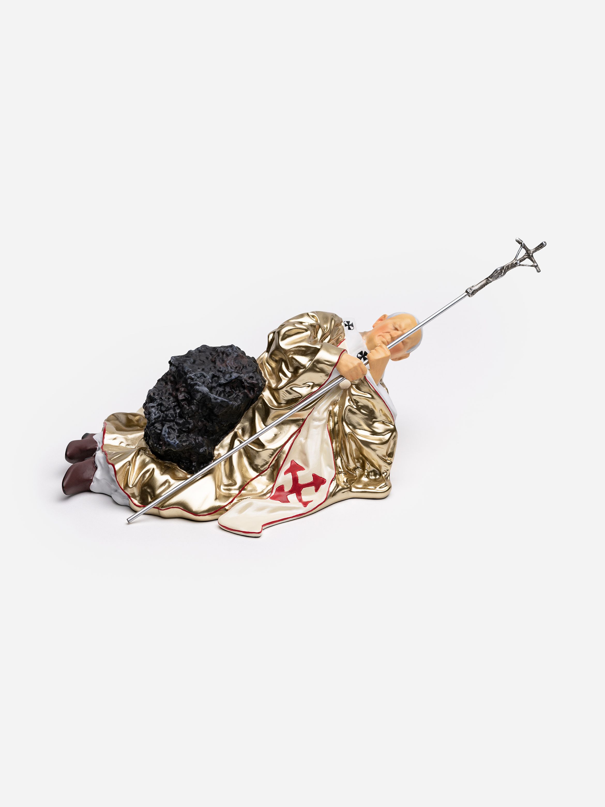 A figurine of a person in a gold robe lying on the ground, holding a long staff with a cross.