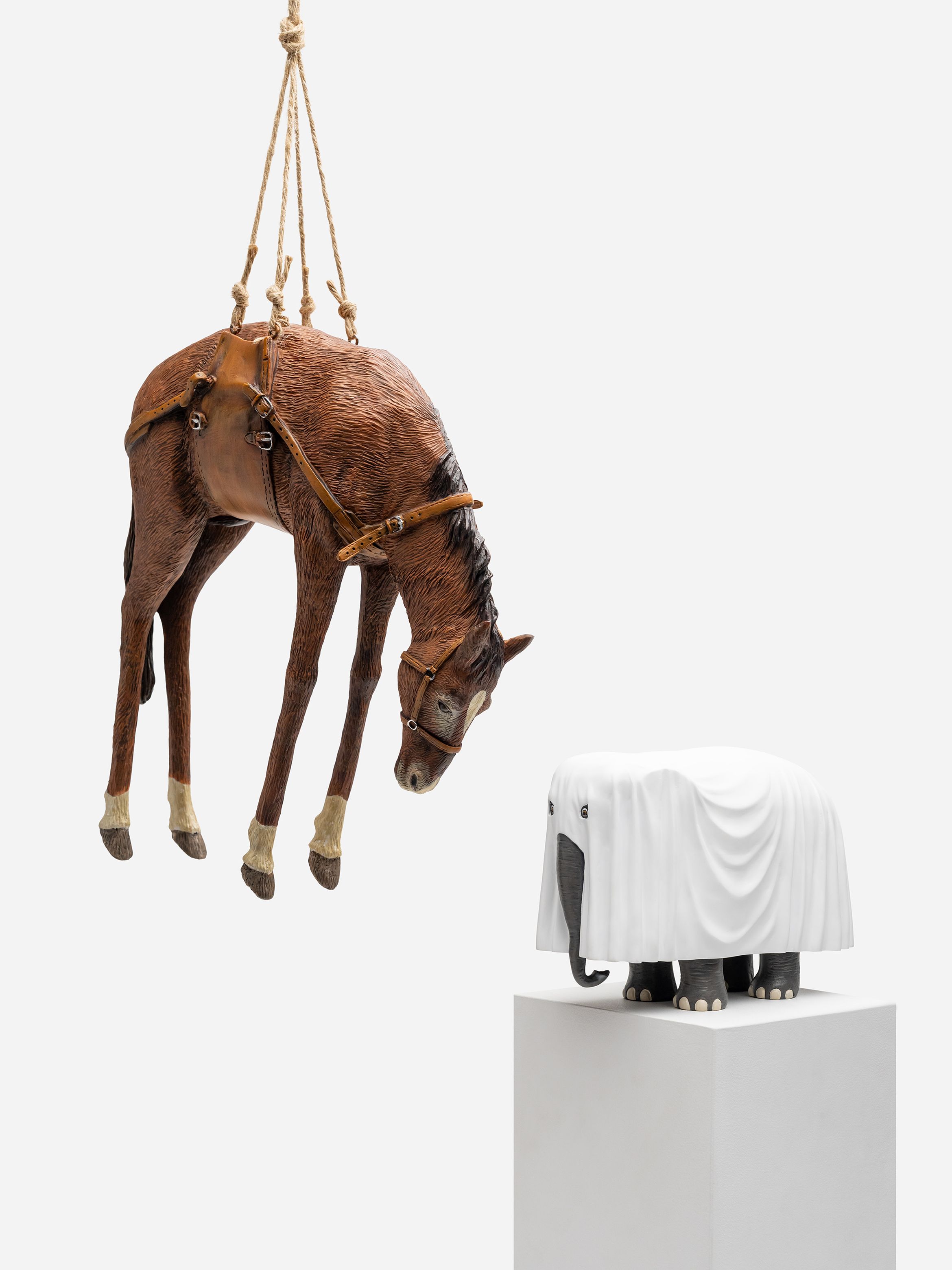 A lifelike horse sculpture is suspended by ropes, facing a smaller sculpture of an elephant covered in a white sheet, both displayed against a white background.