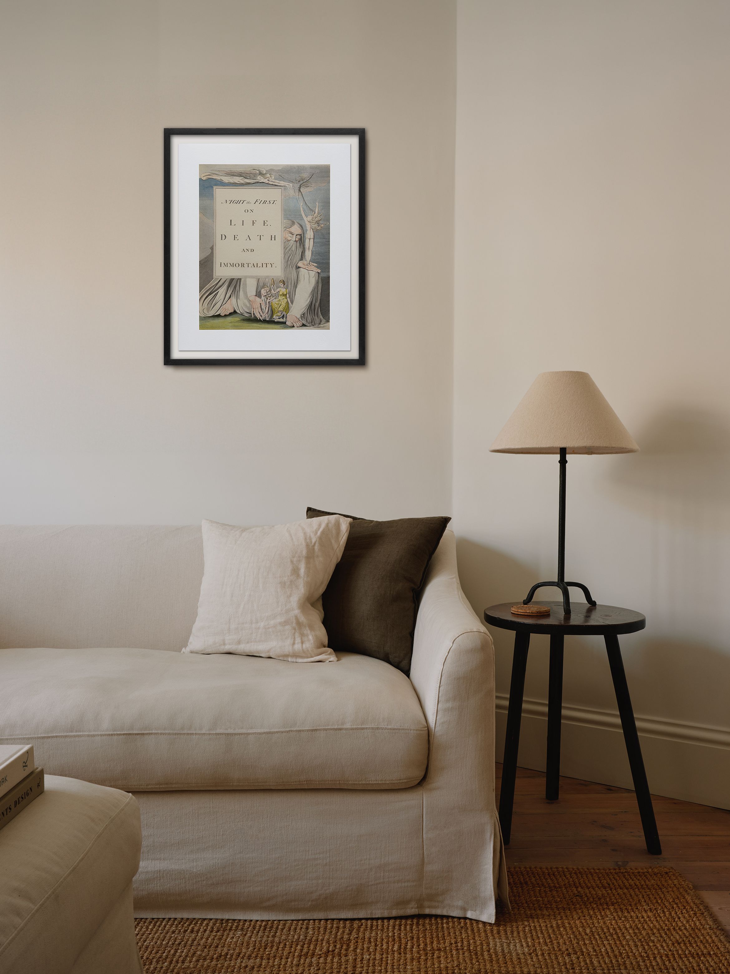 A cosy living room with a beige sofa, two cushions, a side table with a lamp, and a framed artwork on a light-coloured wall.