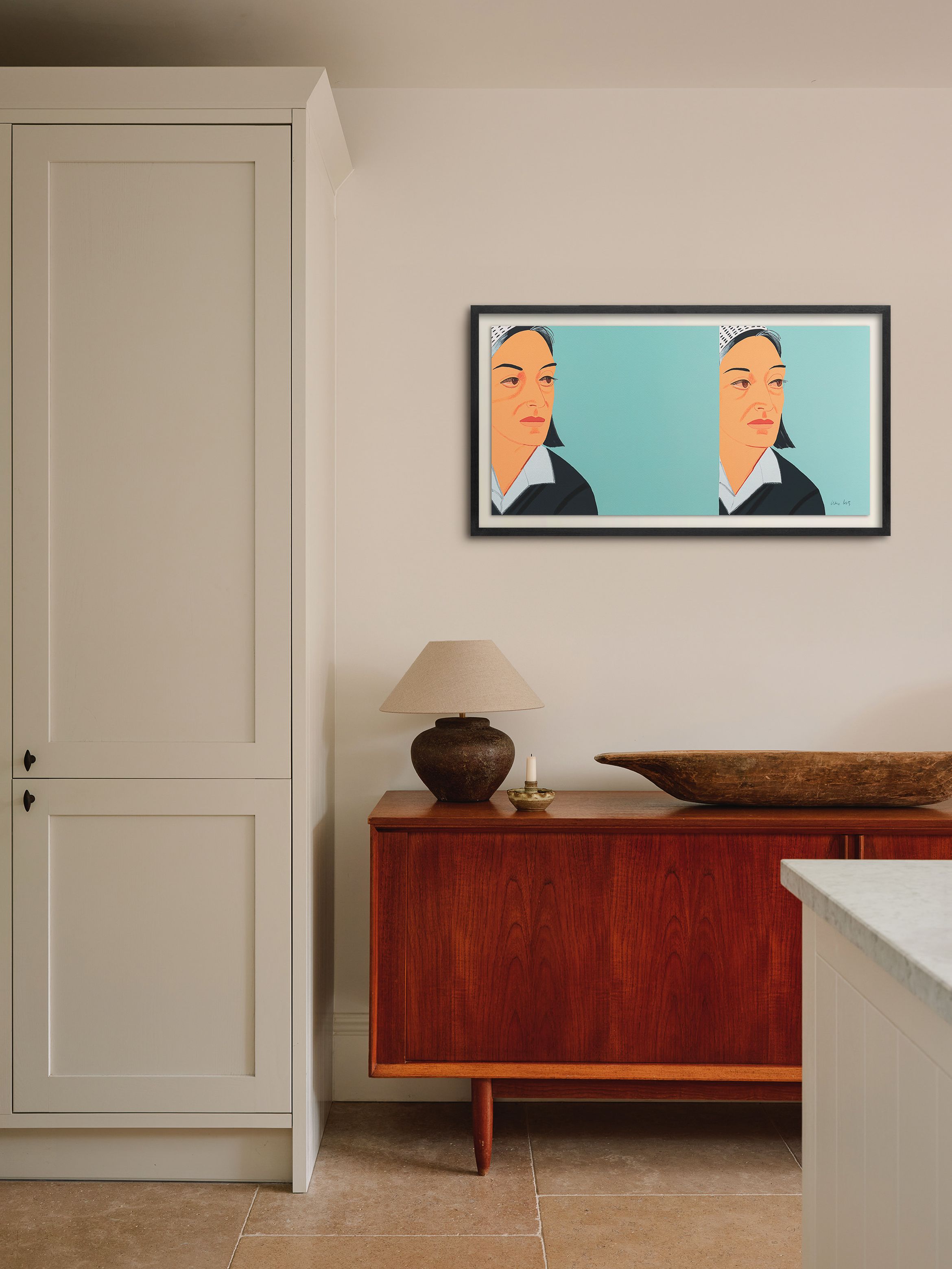 A room with a wooden sideboard, lamp, and decorative bowl. A framed artwork of a woman hangs on the wall above.