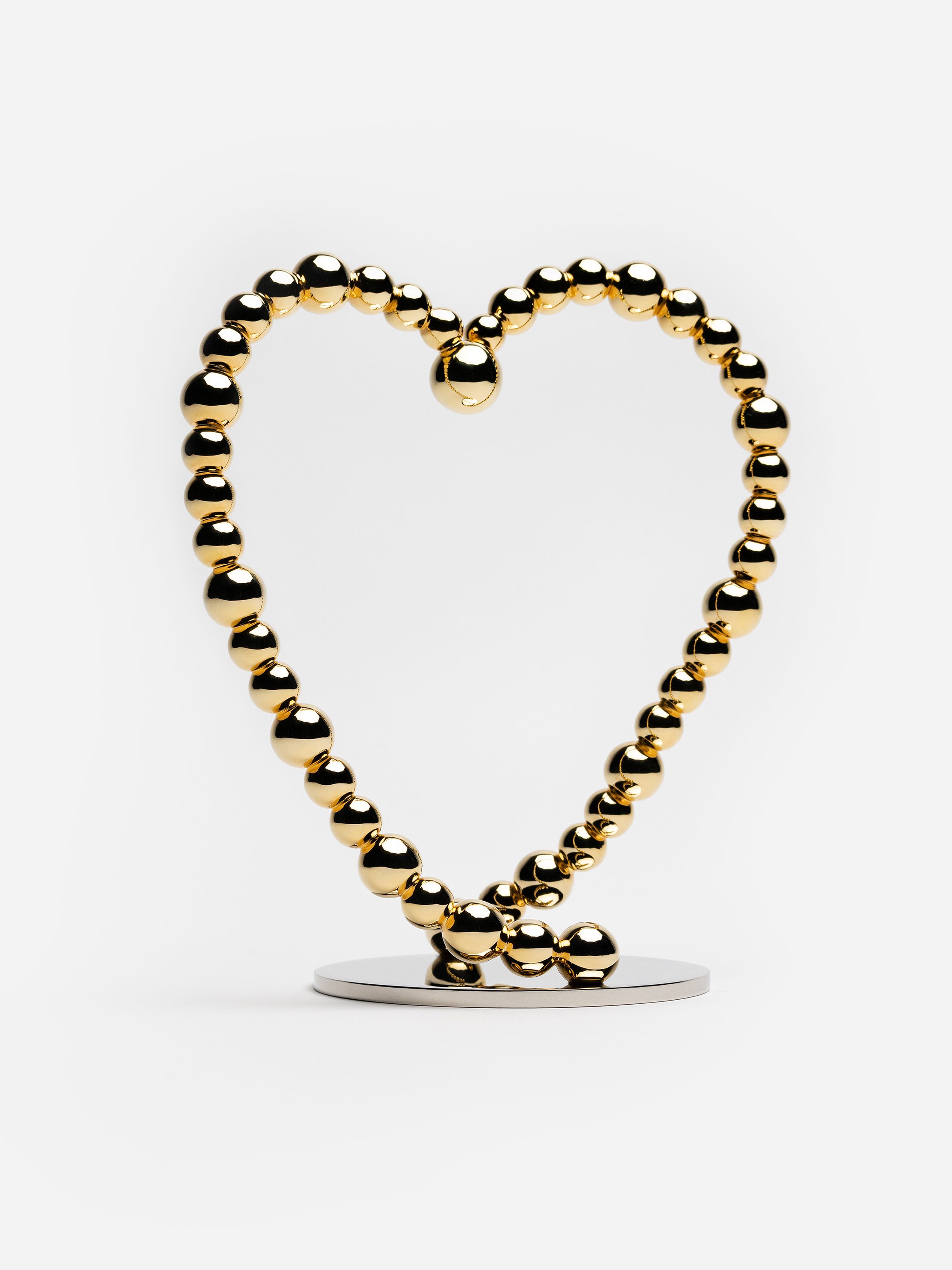 Reflective gold spheres form a heart-shaped sculpture on a round metal base against a white background.