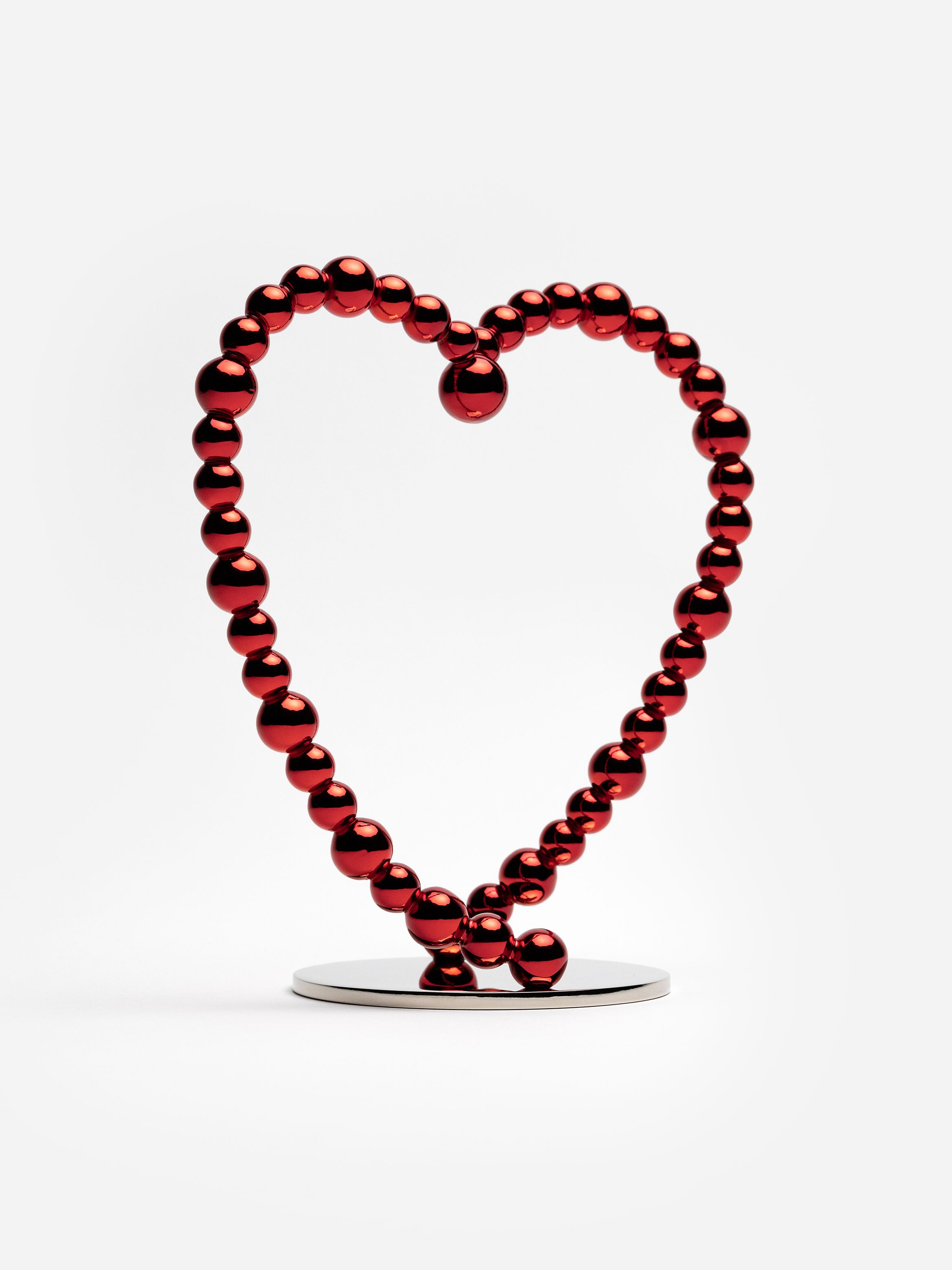 Reflective red spheres form a heart-shaped sculpture on a round metal base against a white background.