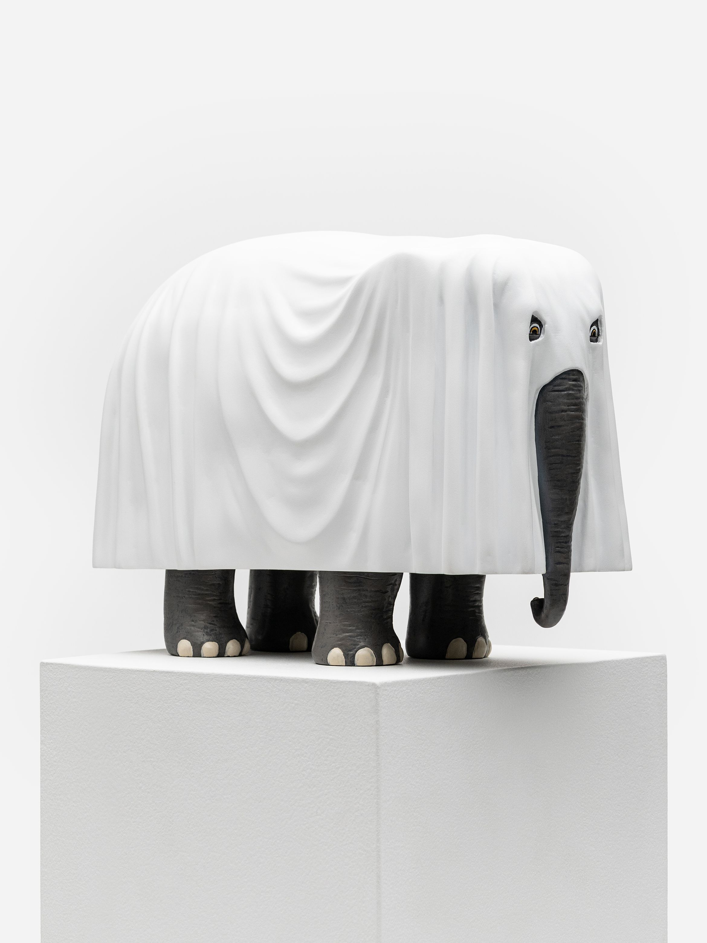 A sculpture of an elephant draped in a white cloth, with grey legs and trunk, standing on a white pedestal. A sculpture of an elephant draped in a white cloth, with grey legs and trunk, standing on a white pedestal.