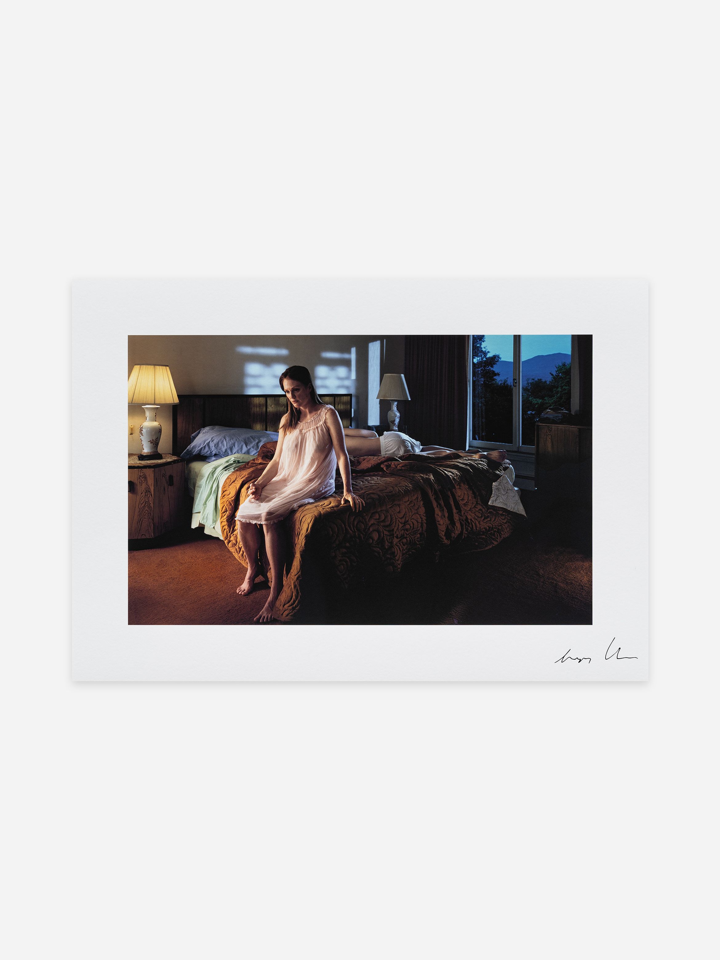 A woman in a nightgown sits on a dimly lit bed in a room with lamps, patterned bedspread, and a window revealing a twilight landscape.