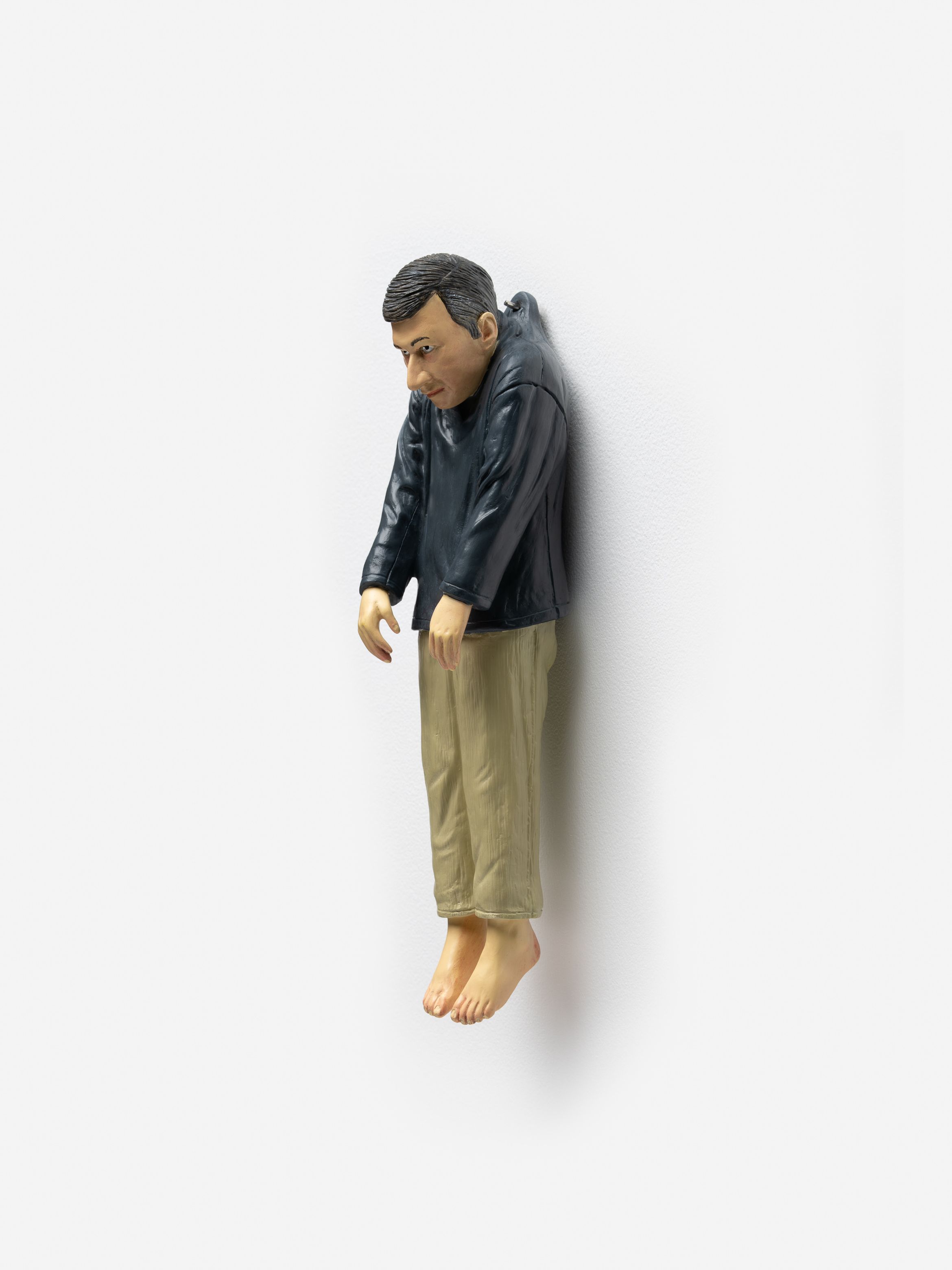 Sculpture of a man in a black jacket and beige pants, hanging limply against a white wall, with bare feet.