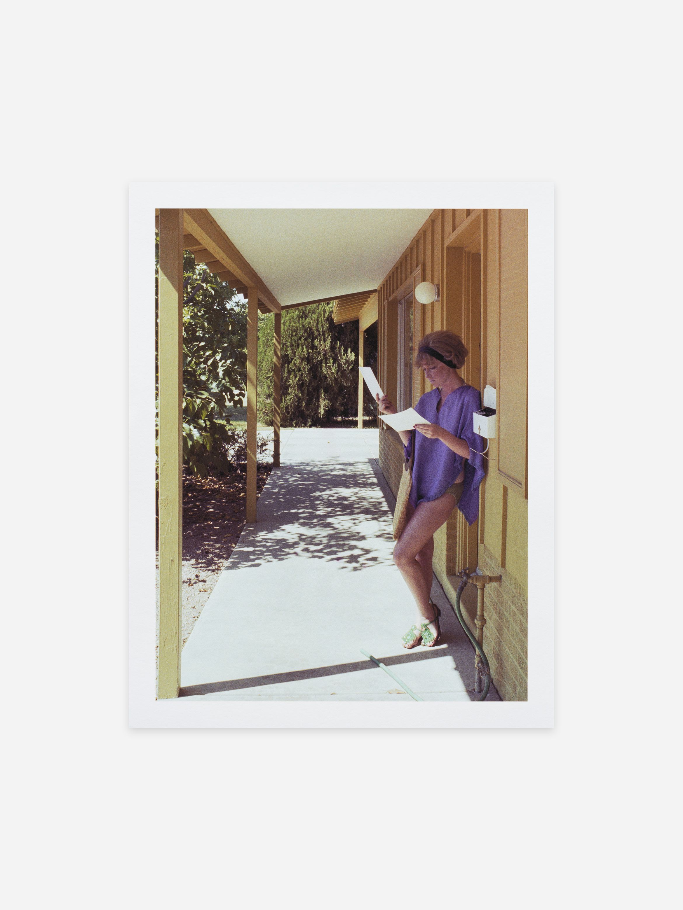 Person in a purple outfit stands under a porch, reading documents, next to a yellow building with trees in the background.