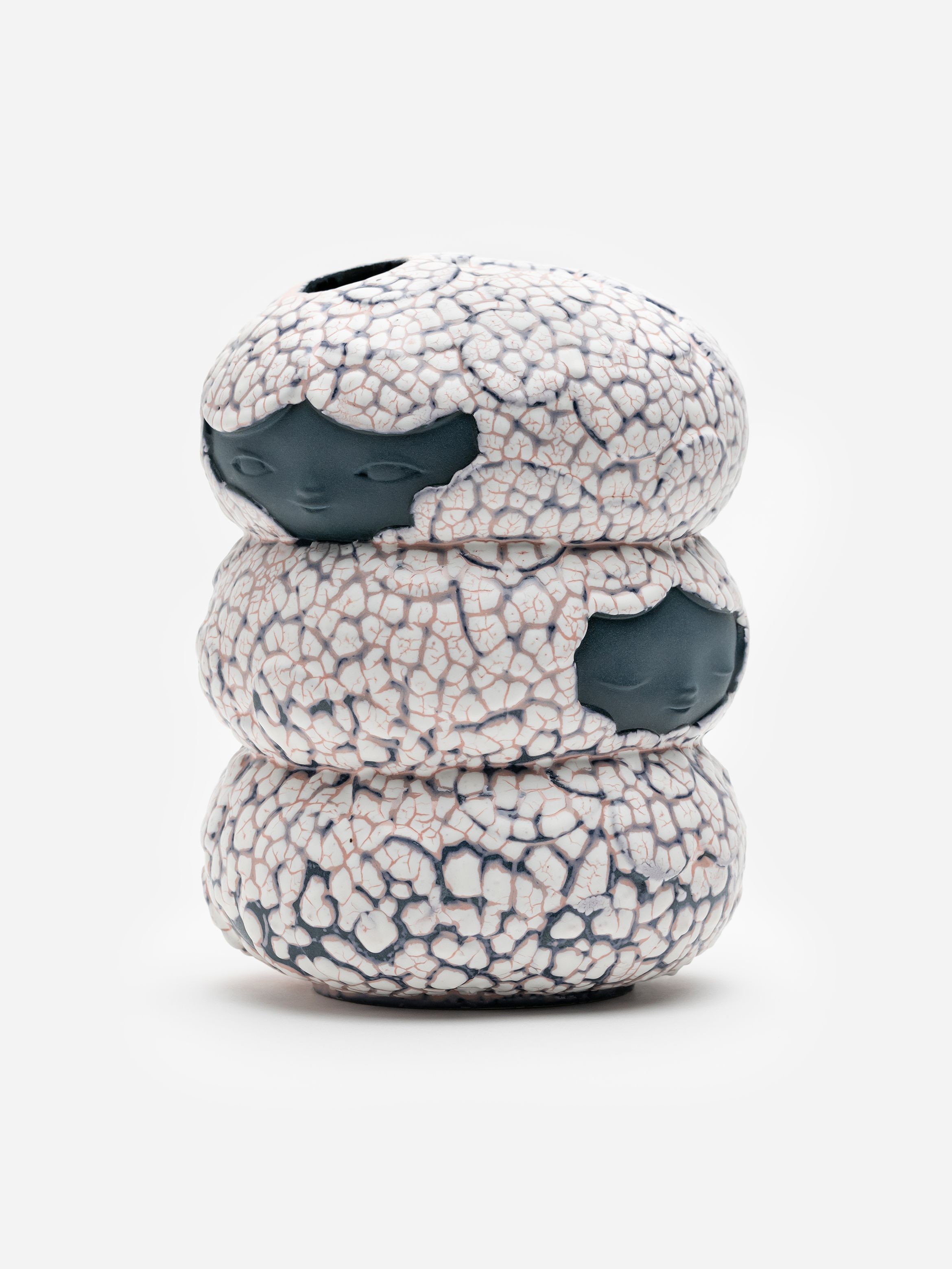 A sculptural object featuring three stacked, textured forms resembling cracked surfaces with subtle facial features on each layer. A sculptural object featuring three stacked, textured forms resembling cracked surfaces with subtle facial features on each layer.
