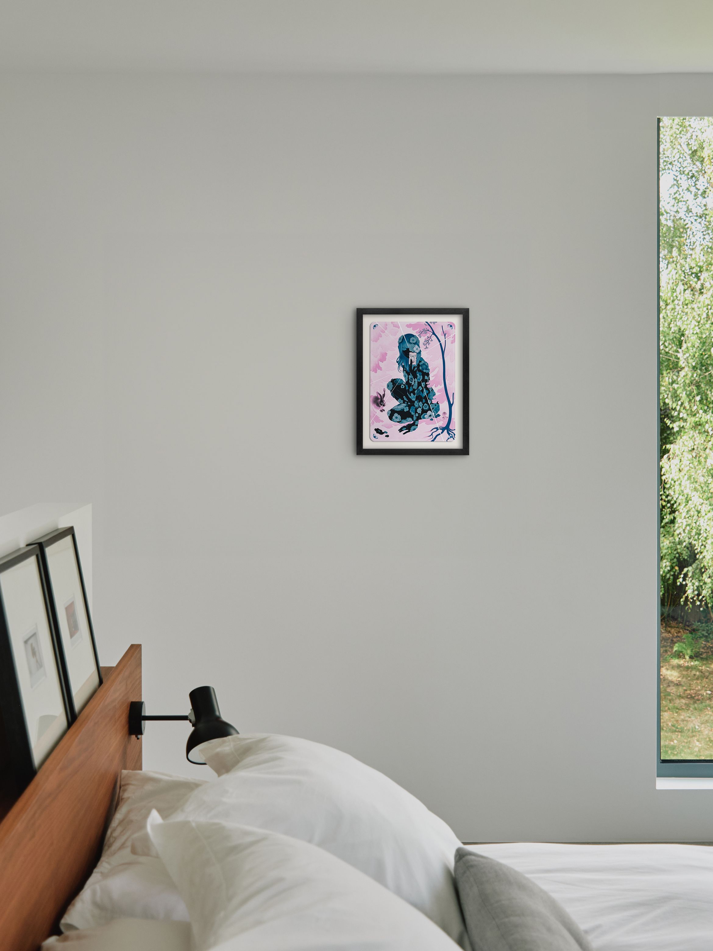 Bedroom with white walls, a bed with white linens, a small framed abstract painting, and a large window showing greenery outside.
