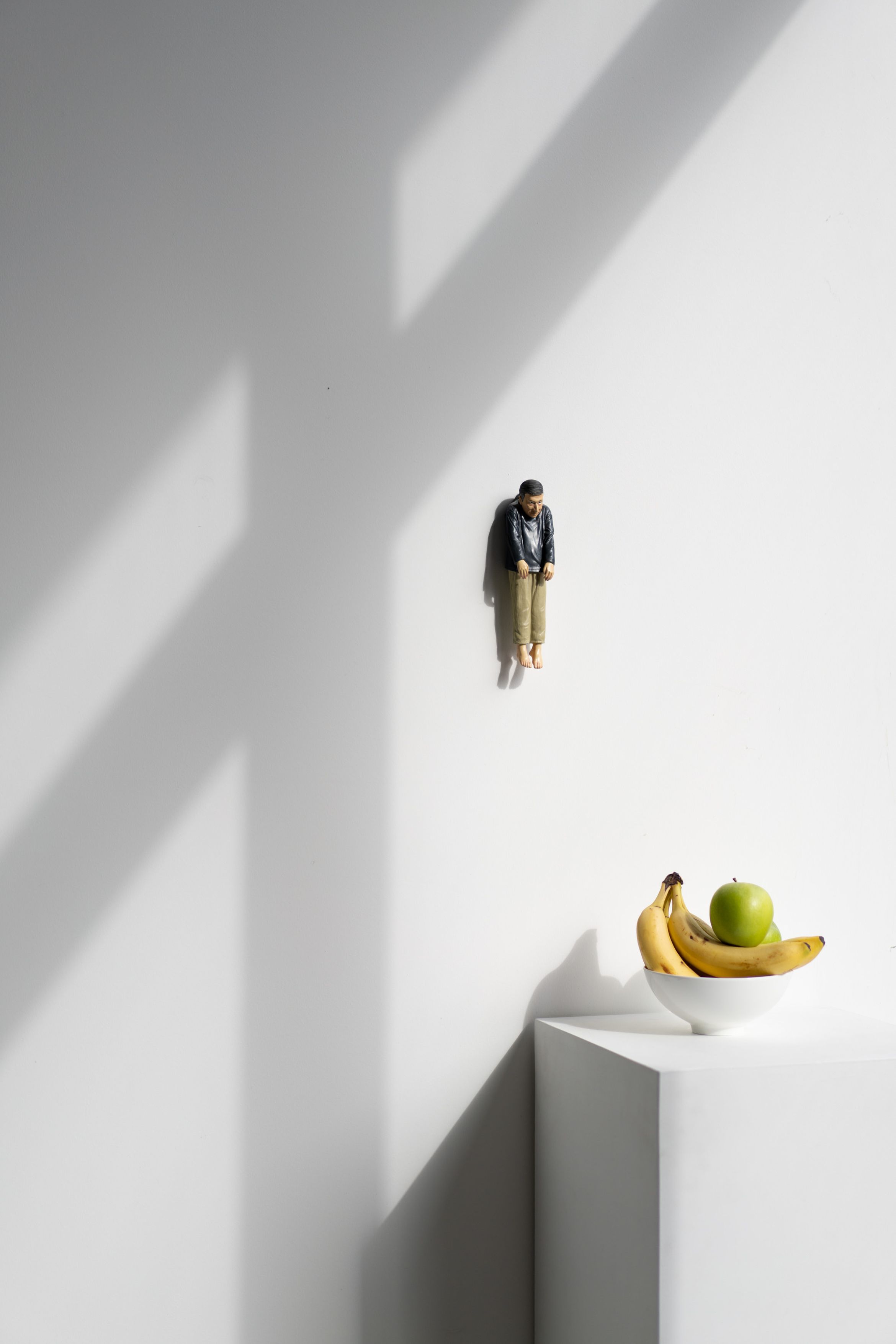 A small figurine on a white wall above a minimalistic white table with a bowl containing bananas and a green apple. Soft shadows cast across the scene.