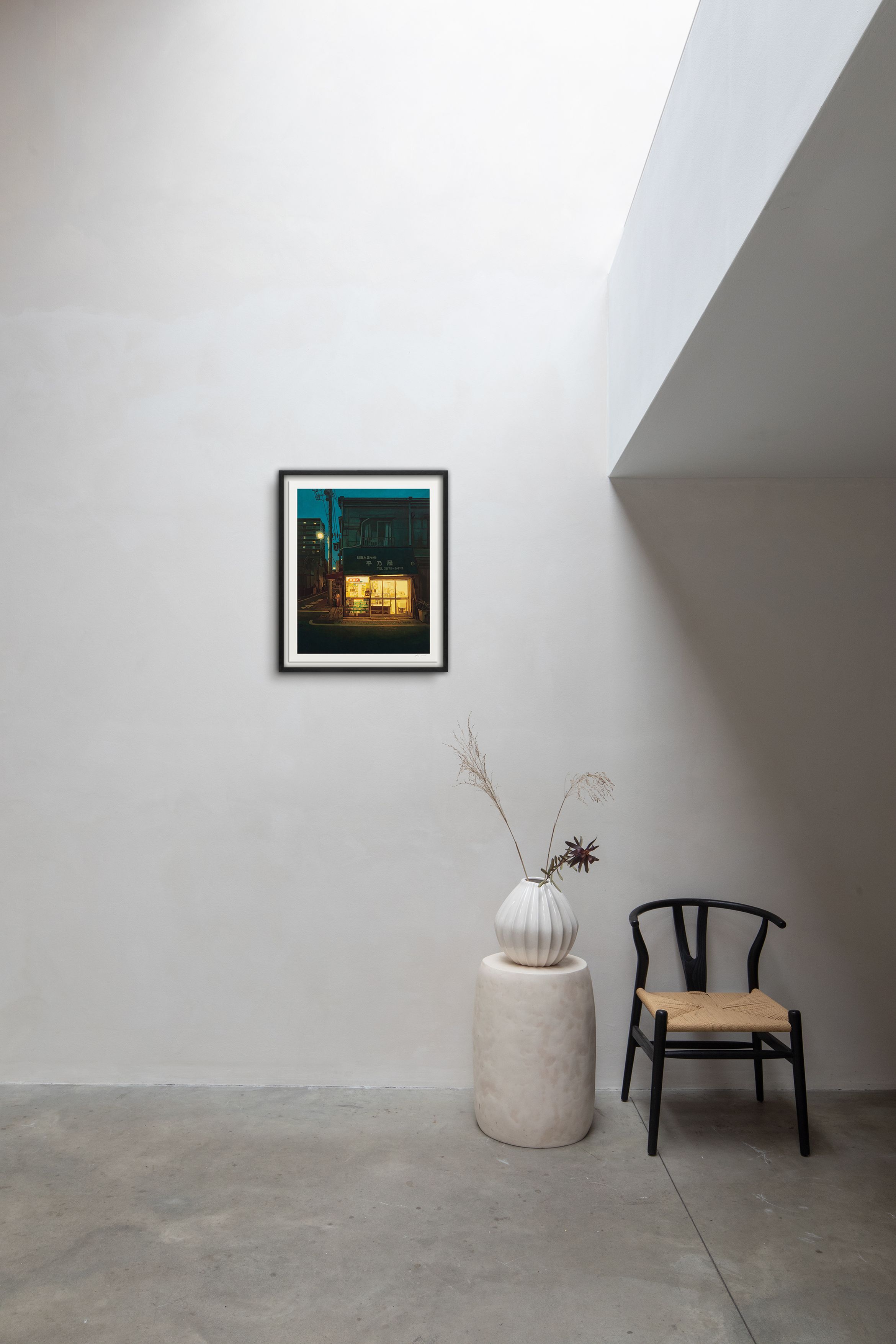 Minimalist room with a framed picture on a white wall, a black chair, and a vase with dried flowers on a round pedestal.