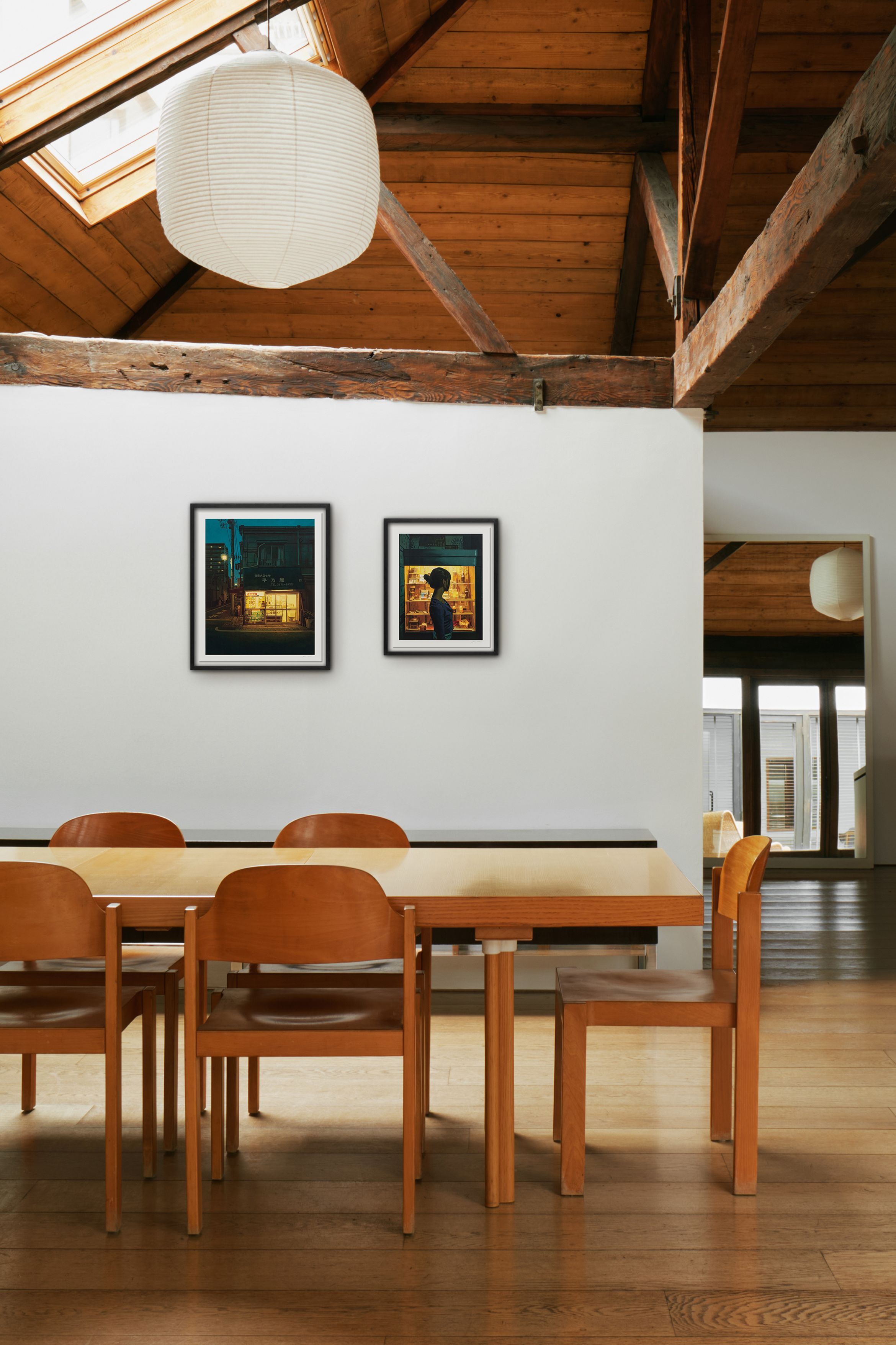 Dining area with a wooden table and chairs, white walls, framed art, exposed beams, and a paper lantern under a skylight.