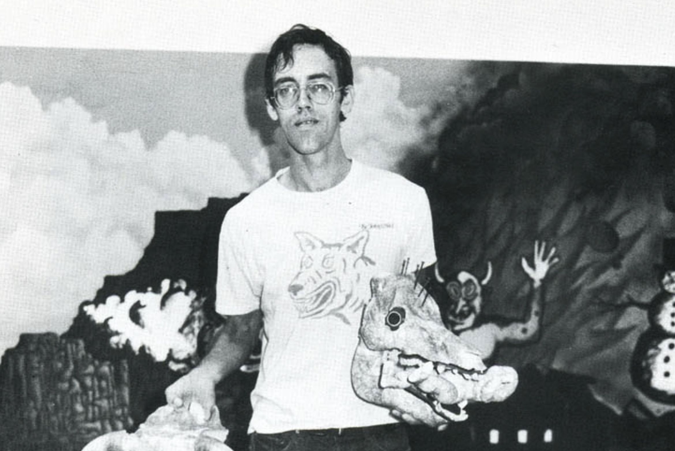 A man with glasses holds a carved animal head sculpture, standing in front of a mural featuring clouds and mythical figures.