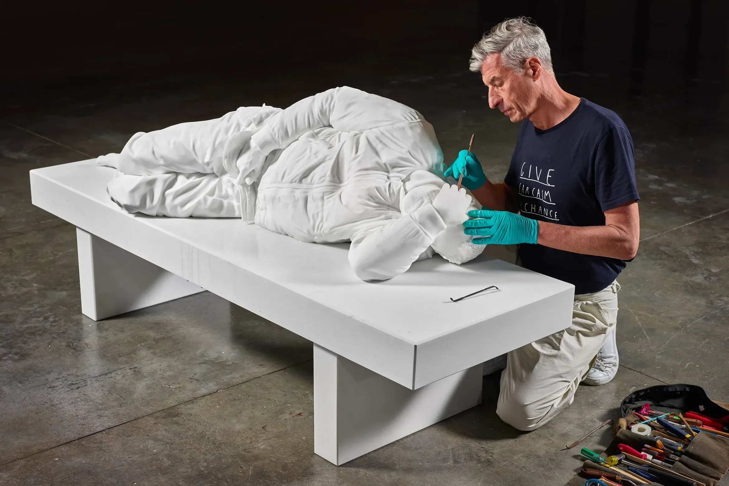 Maurizio Cattelan works on a sculpture of a man laying down on a bench