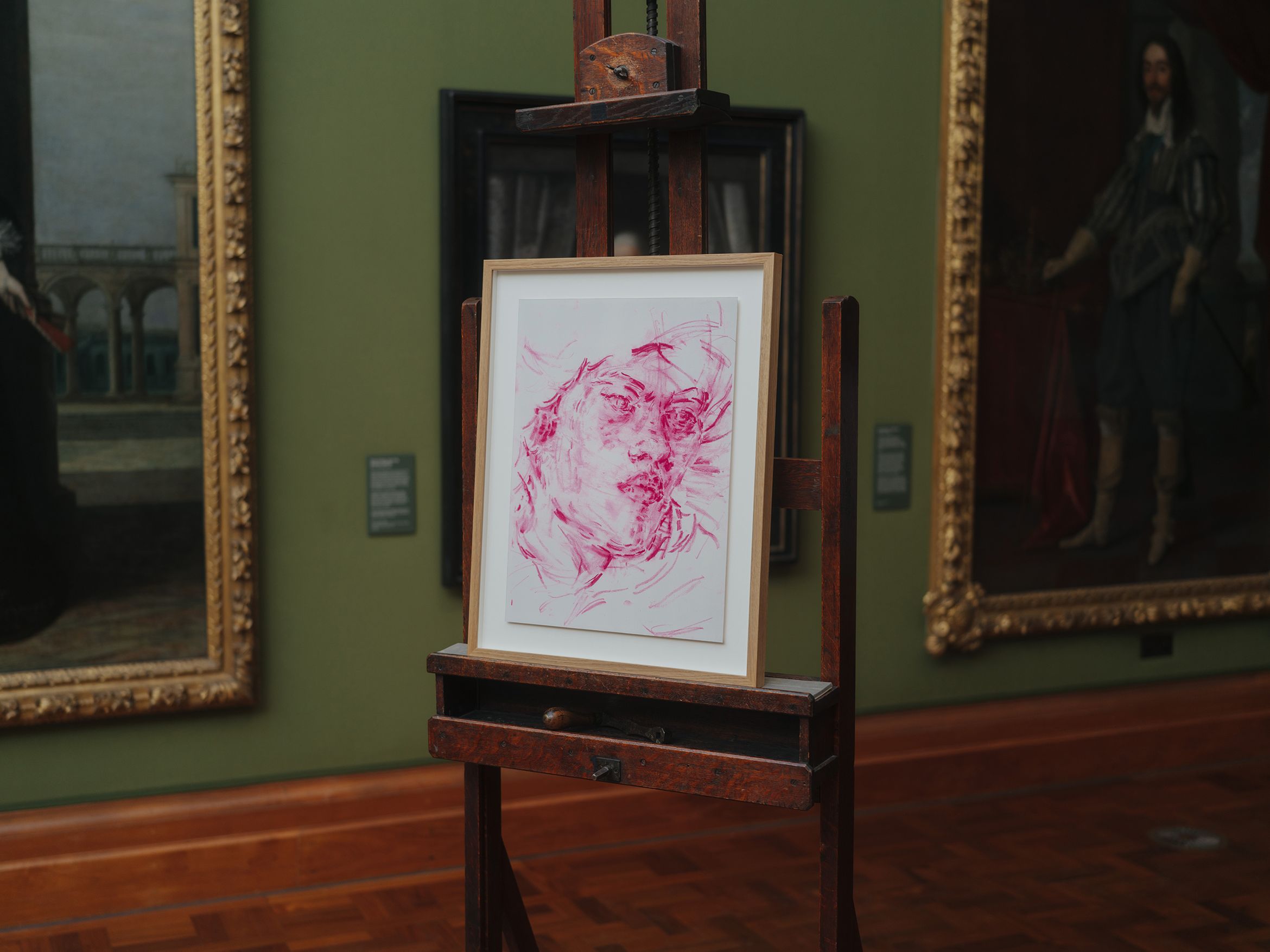 A framed print of a face in red tones on an easel, displayed in an art gallery with paintings on the green walls.