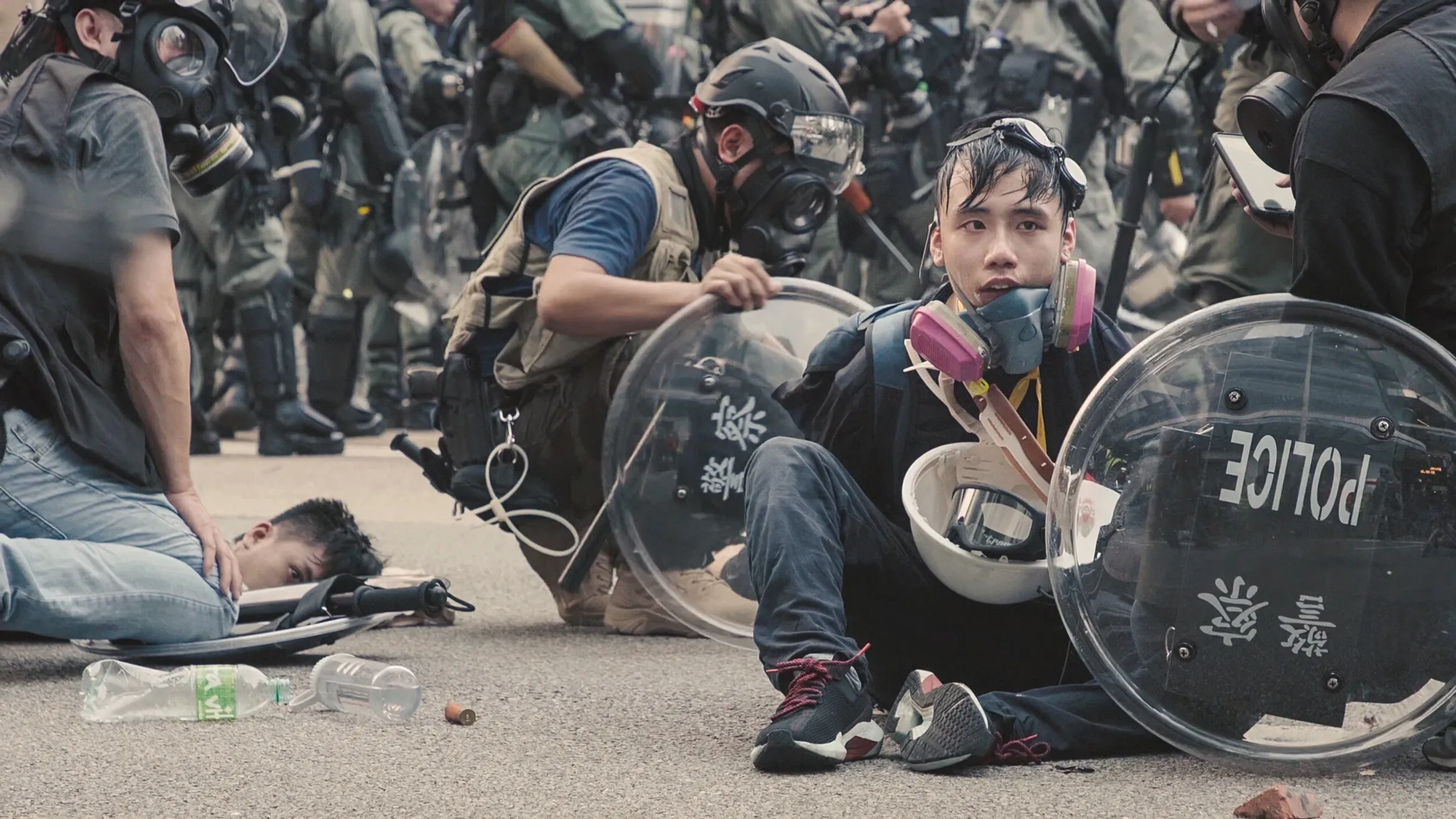 Protesters and police in riot gear clash; one protester sits on the ground with a shield, wearing a gas mask, surrounded by debris.