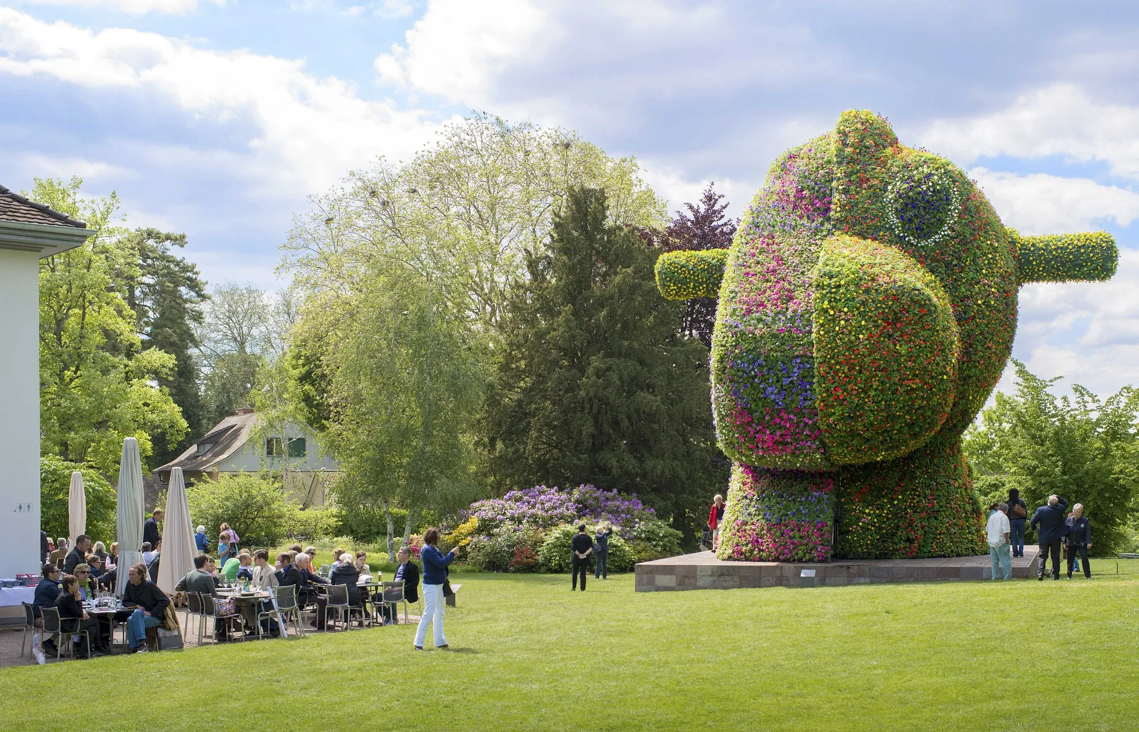 Large colorful floral sculpture of a cartoonish dog in a park; people dining and strolling nearby.