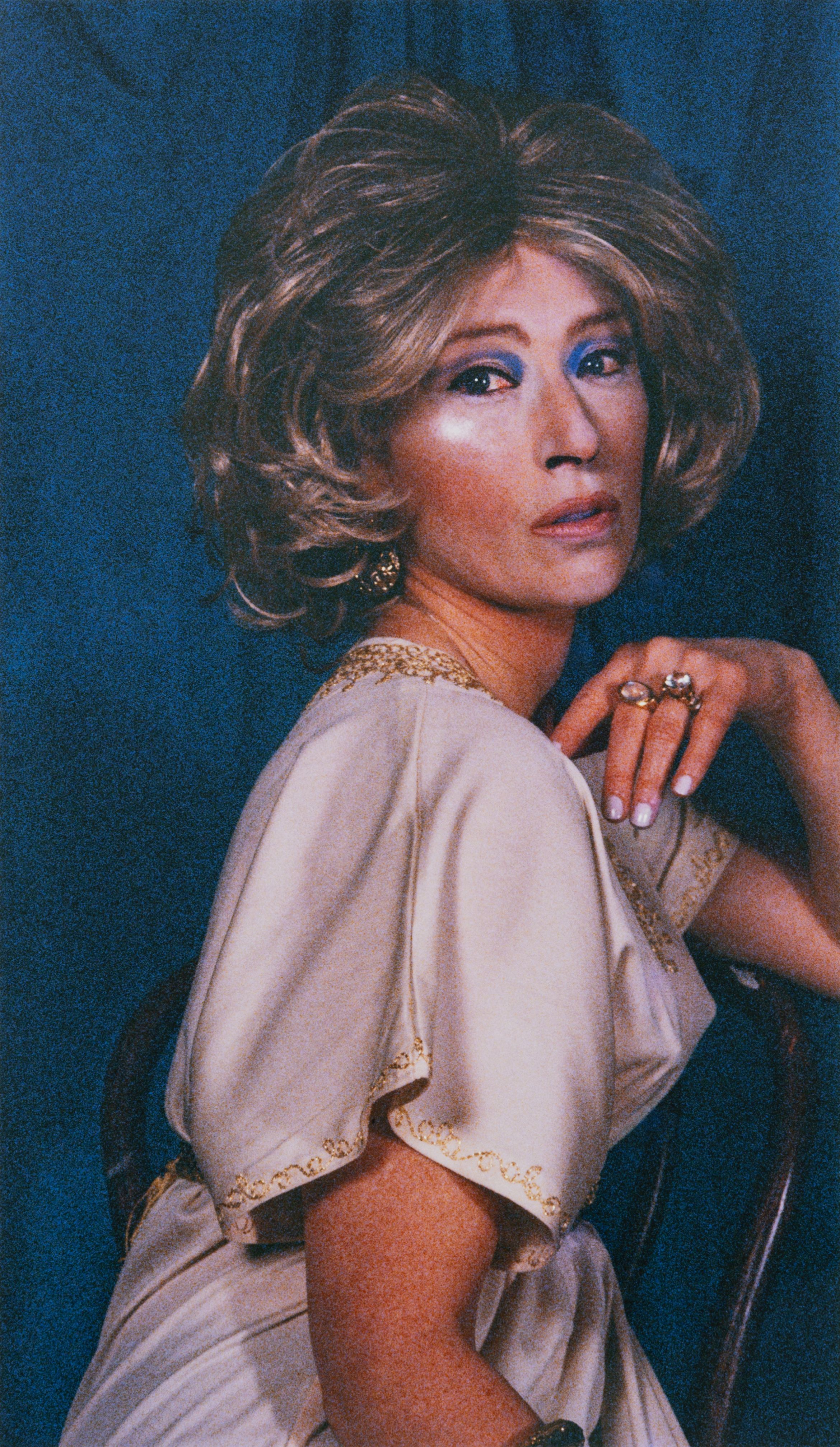 Person in dress and make-up, shot against a blue backdrop.