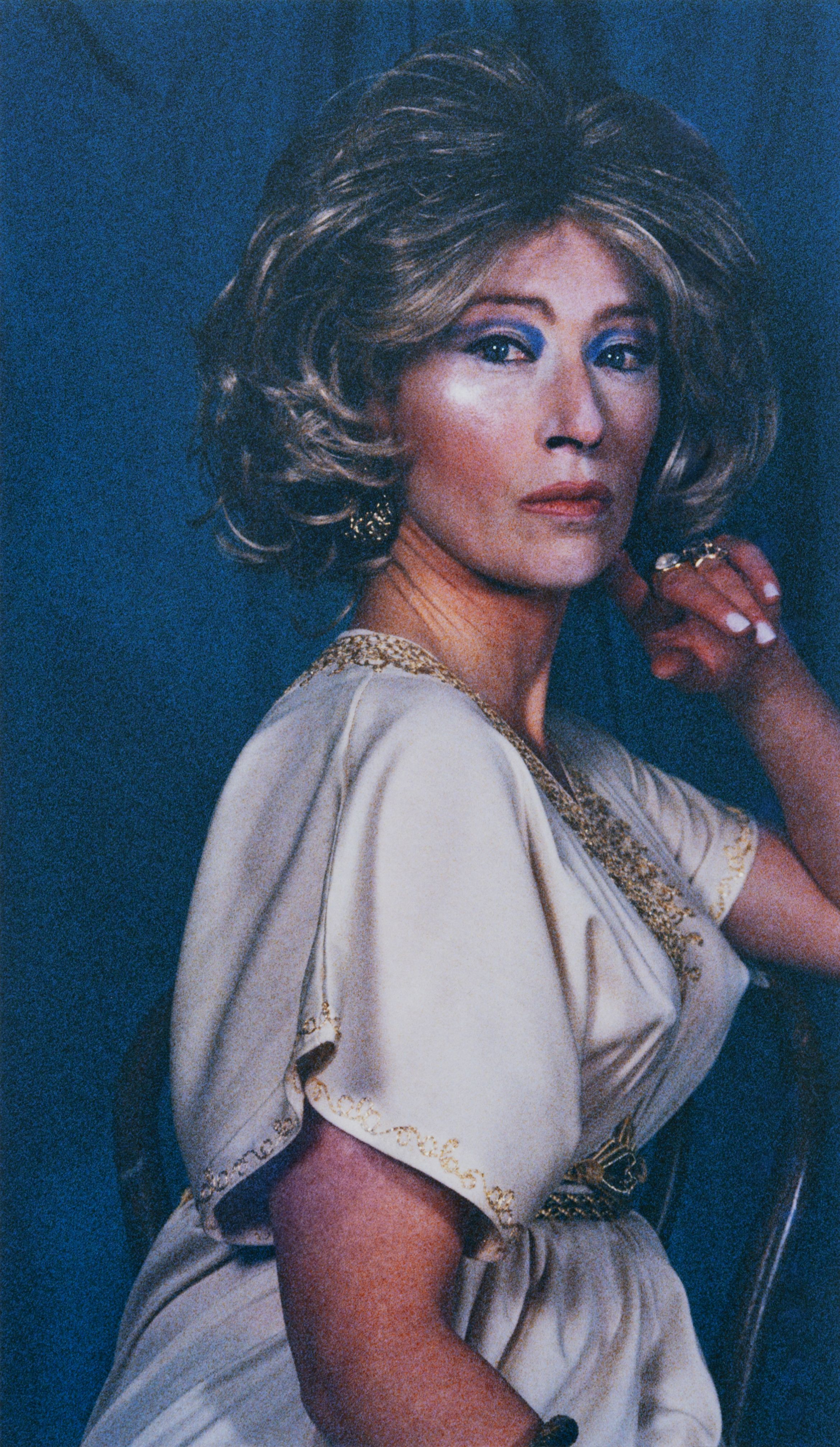 Person in a vintage-style dress and curly wig, sitting against a dark background, gazing thoughtfully with hand near chin.