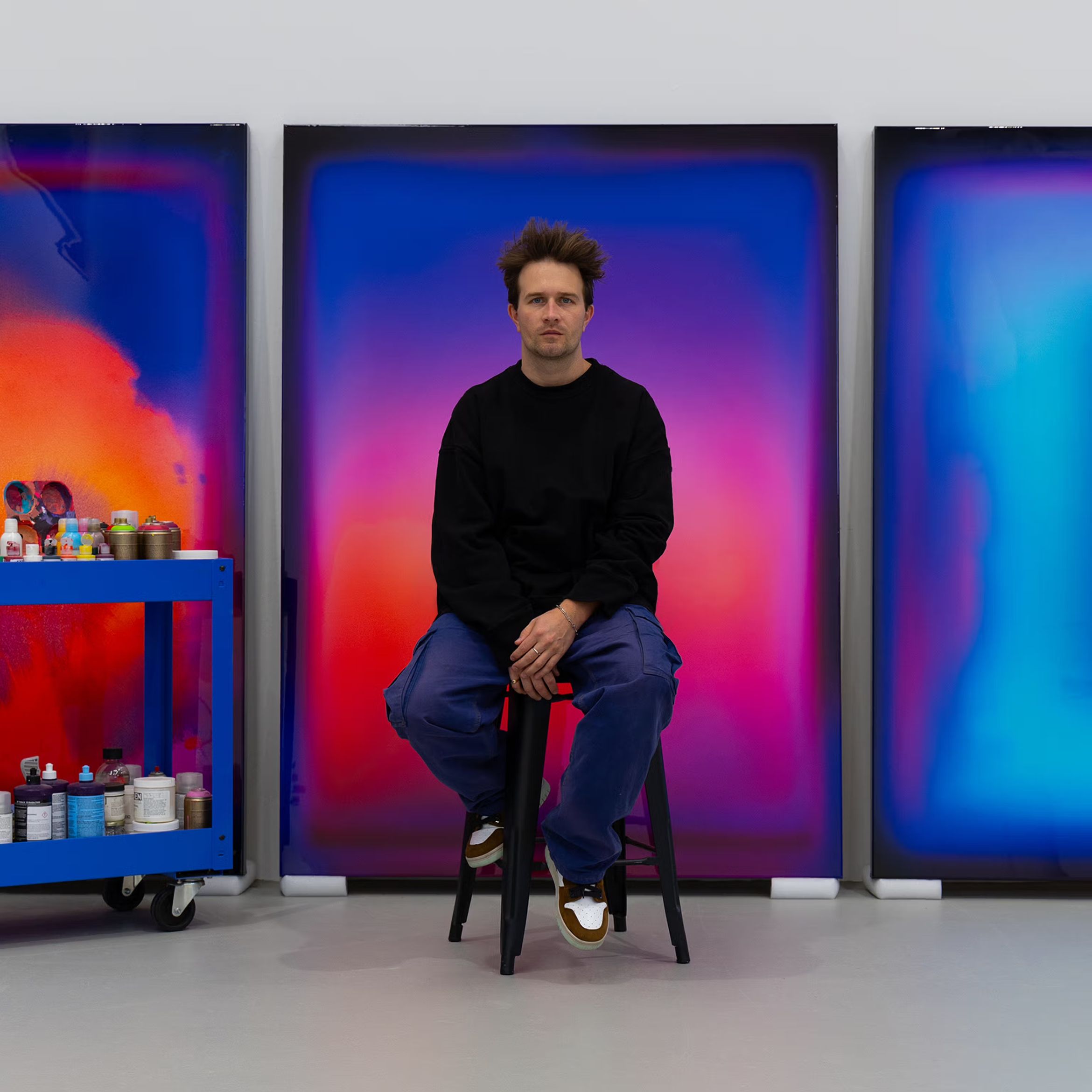 Artist Nick Thomm seated in front of large, vibrant abstract paintings with a cart of paints on the side in a studio setting.